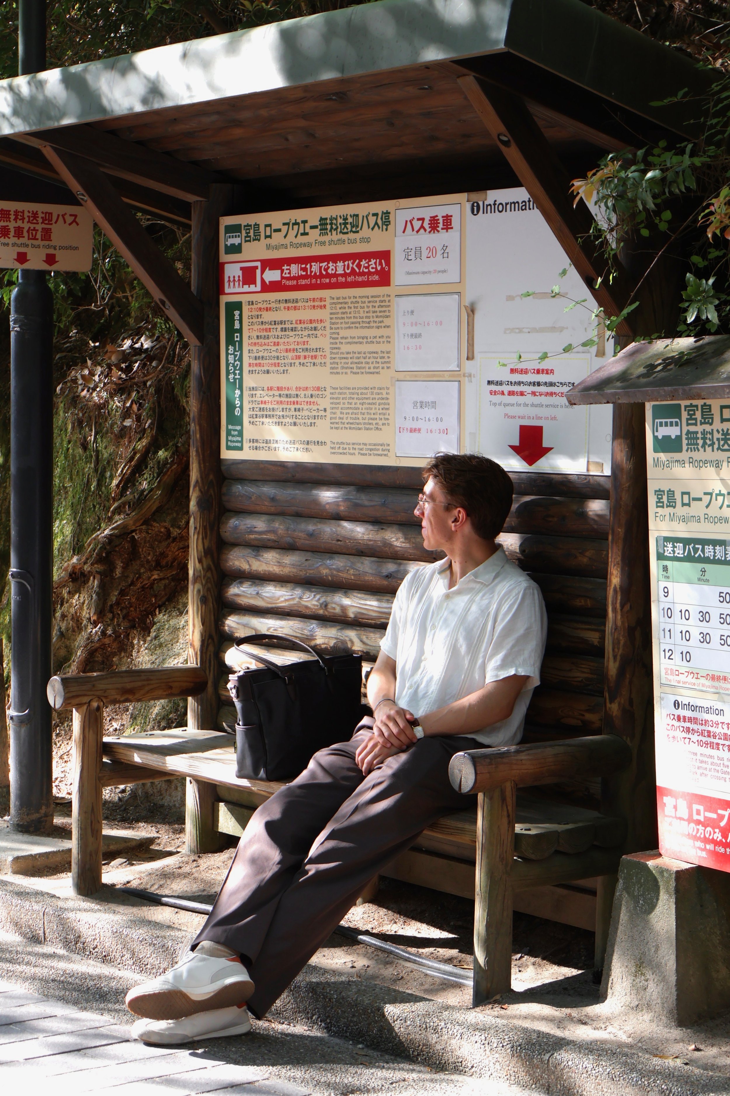 A man sitting on a wooden bench at a bus stop, holding a black bag, with a sign and informational boards behind him, amidst trees and natural surroundings.