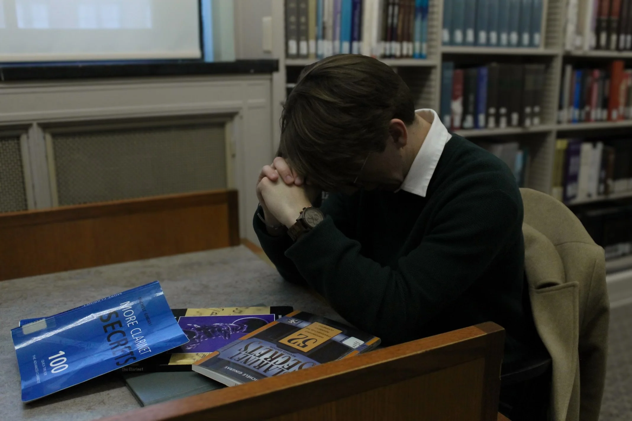 A person with brown hair, wearing a black sweater and a watch, sitting at a table with their head bowed and hands clasped. There are books and pamphlets on the table, and a background of bookshelves in a library.