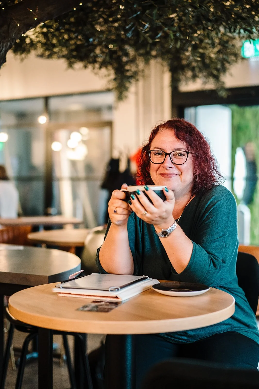 A woman with curly red hair, glasses, and a watch sitting at a wooden table in a cafe, holding a saucer with a cup of coffee and smiling.