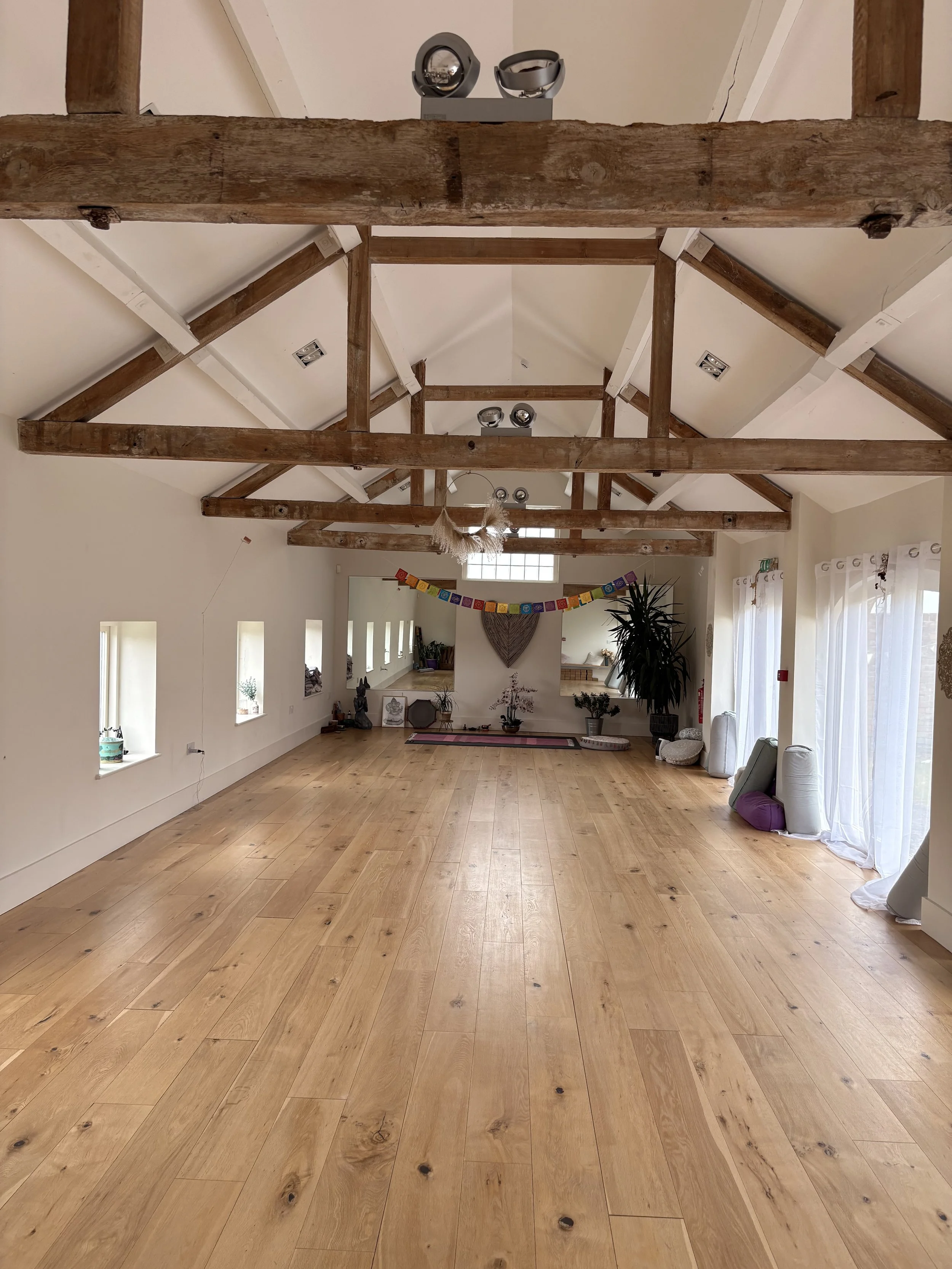 A spacious room with high ceilings and exposed wooden beams. The floor is made of light-colored wood. On the right side, large windows with white curtains let in natural light. Decor include a colorful bunting, potted plants, and various decorative items.