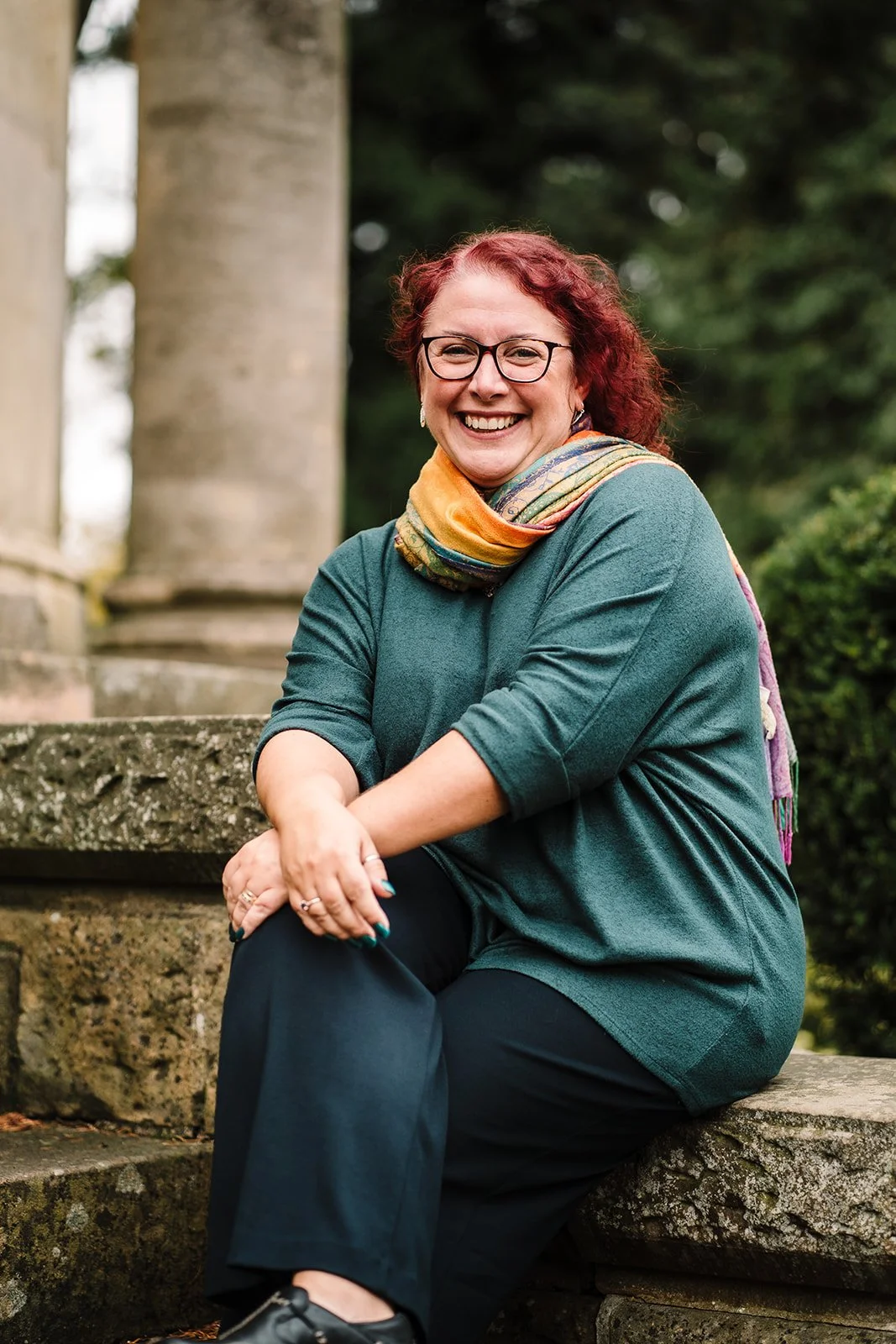 A woman with red hair and glasses smiling while sitting outdoors on a stone bench, wearing a teal top and a colorful scarf, with greenery in the background.