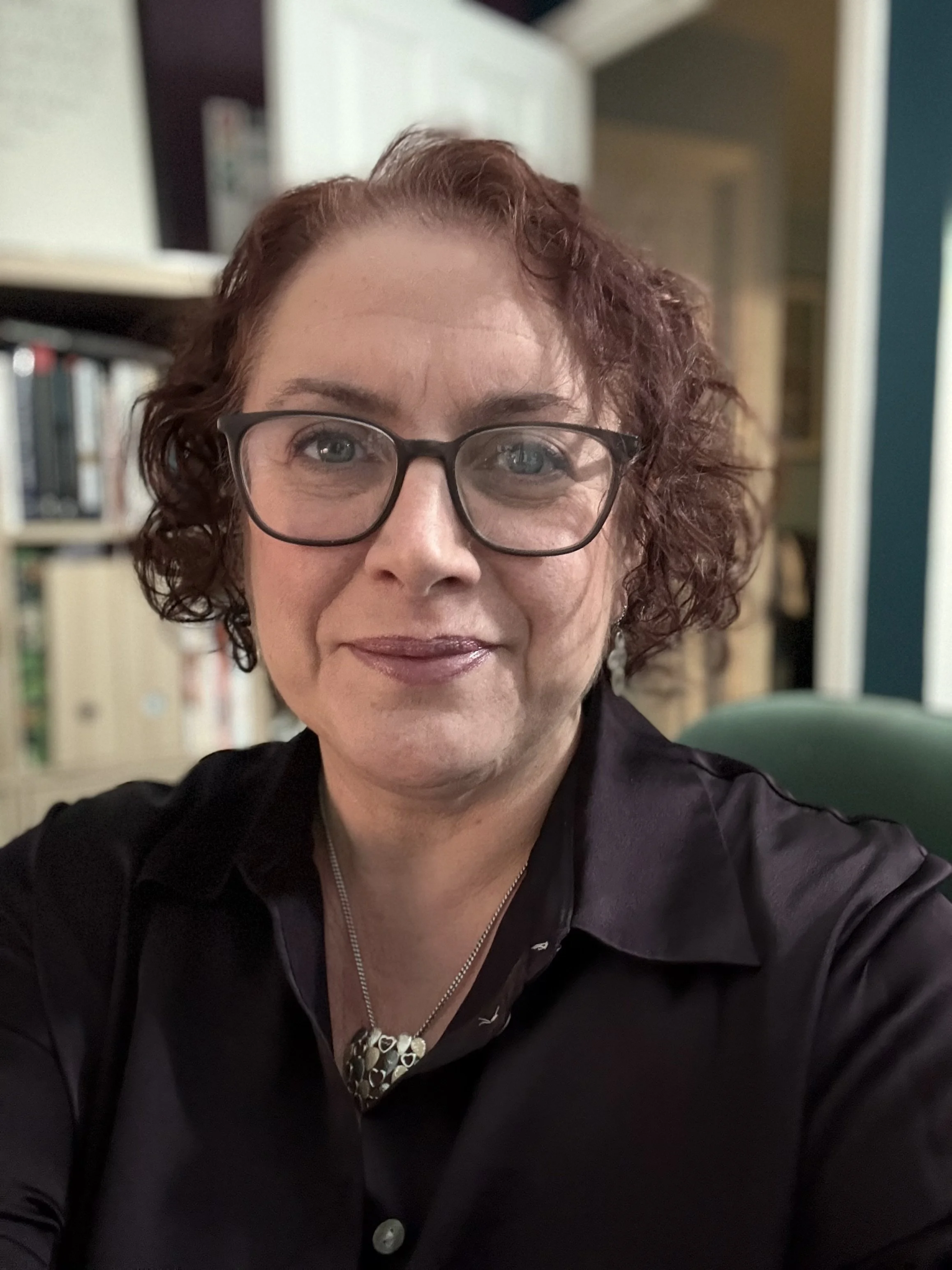 A woman with curly red hair and glasses taking a selfie in a room with bookshelves in the background.
