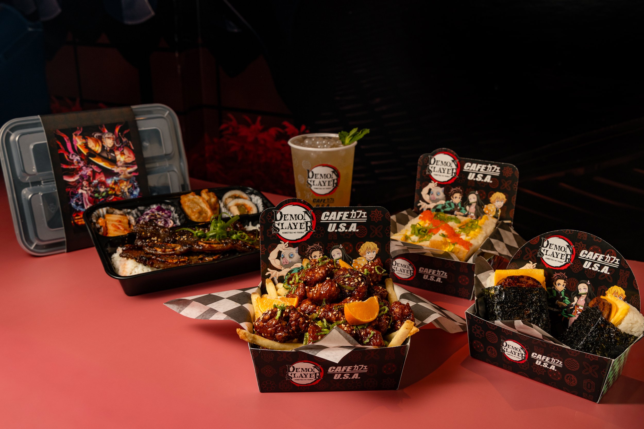 Assorted Japanese-inspired dishes including sushi rolls, fried chicken, rice bowls, and a drink, arranged on a table with Demon Slayer themed food packaging and decorations.