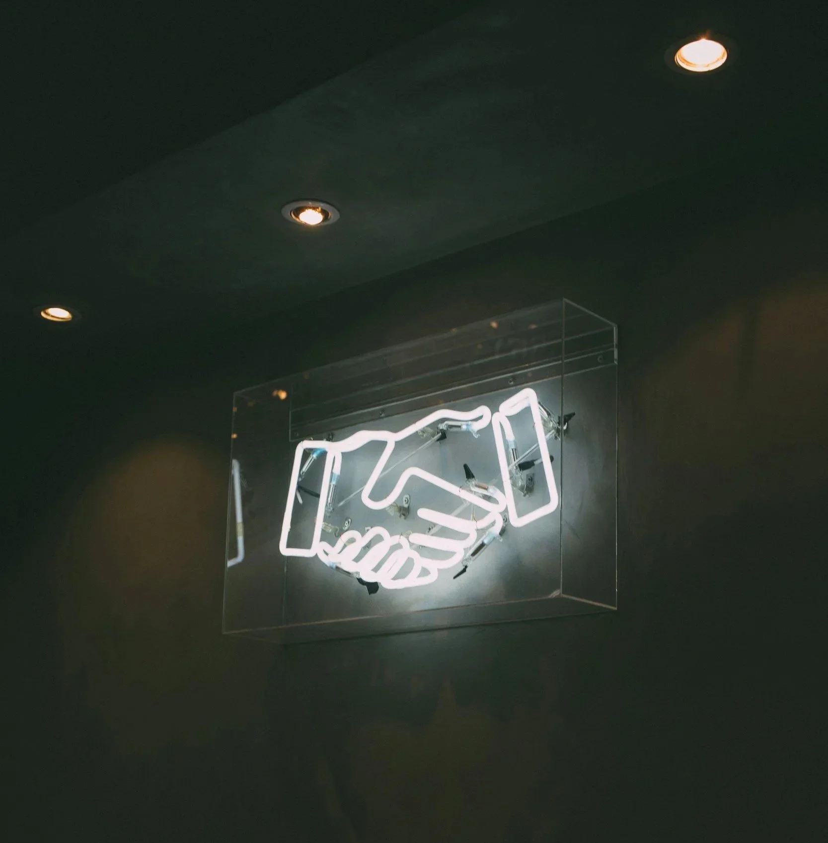 Neon sign of a handshake inside a glass display case, against a dark wall with ceiling lights.