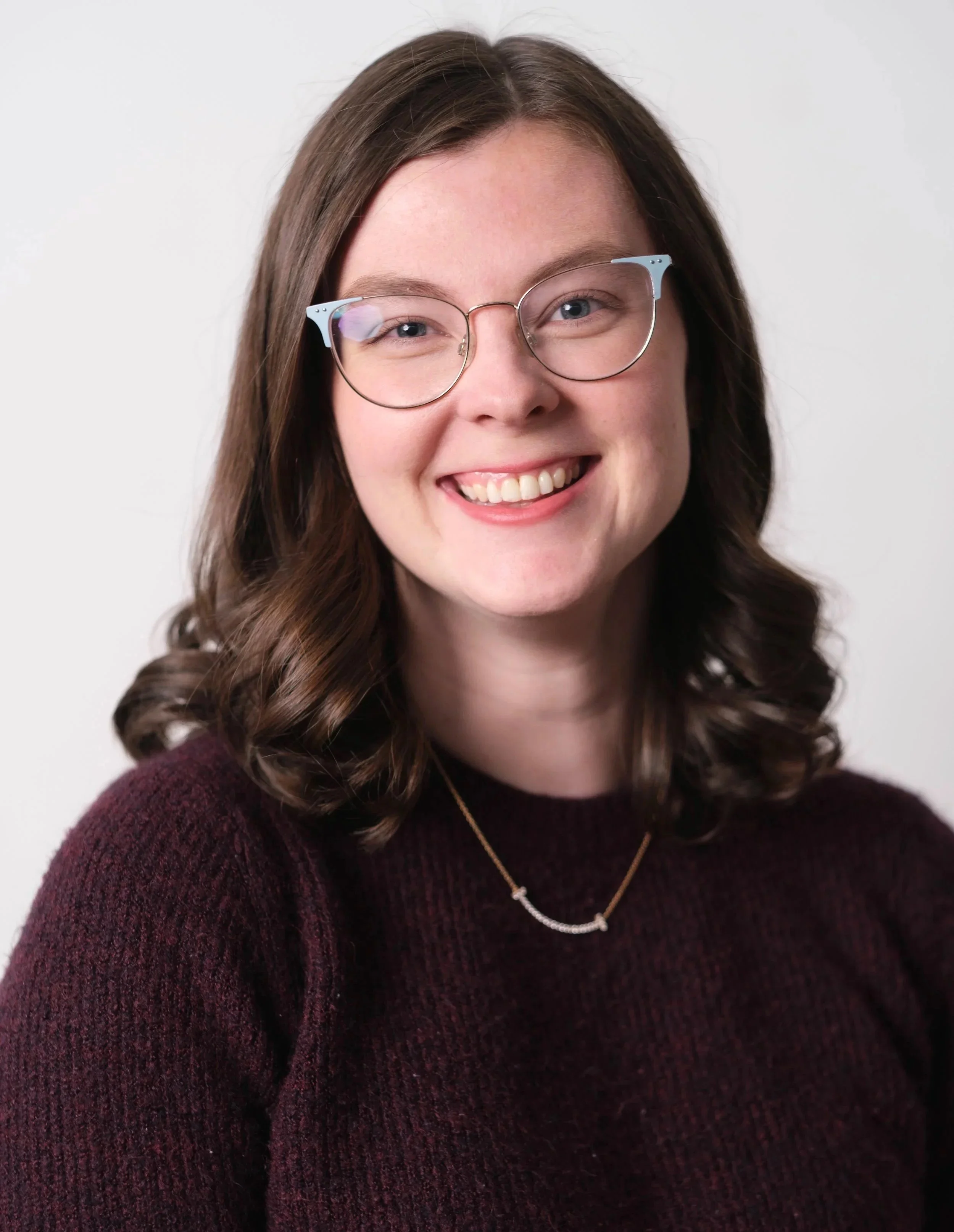 Close-up of a woman with brown wavy hair and glasses, smiling, wearing a maroon sweater and a delicate necklace, against a plain white background.