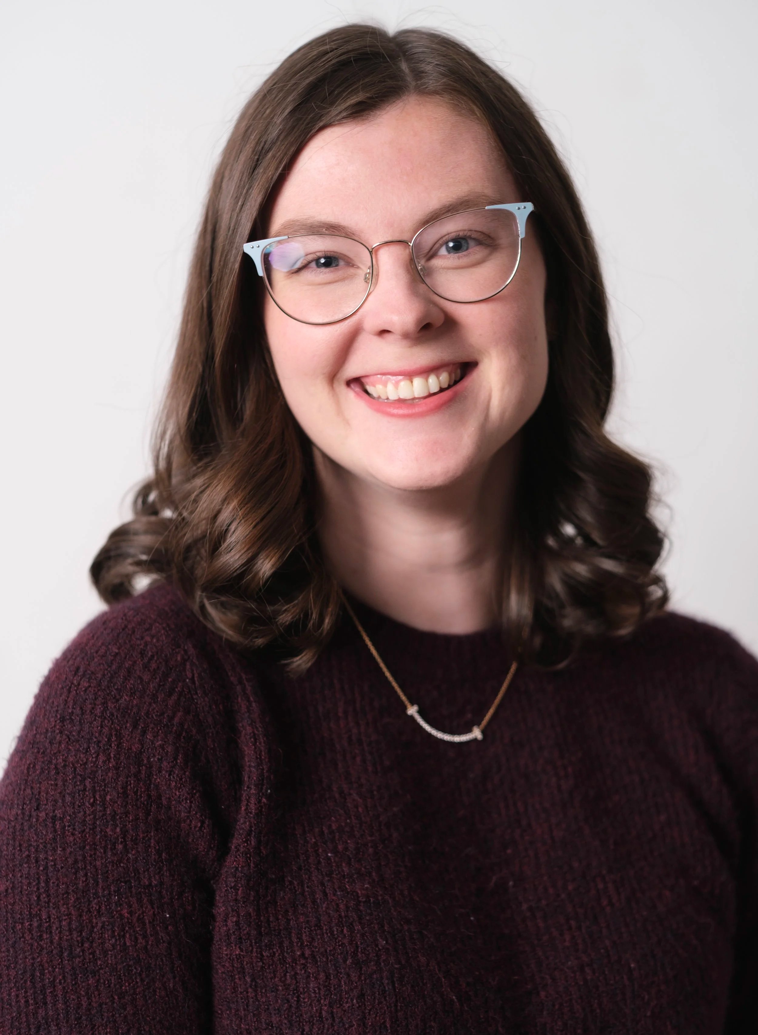 Portrait of a young woman with wavy brown hair, wearing glasses with light blue accents, smiling, and dressed in a burgundy sweater with a gold necklace.