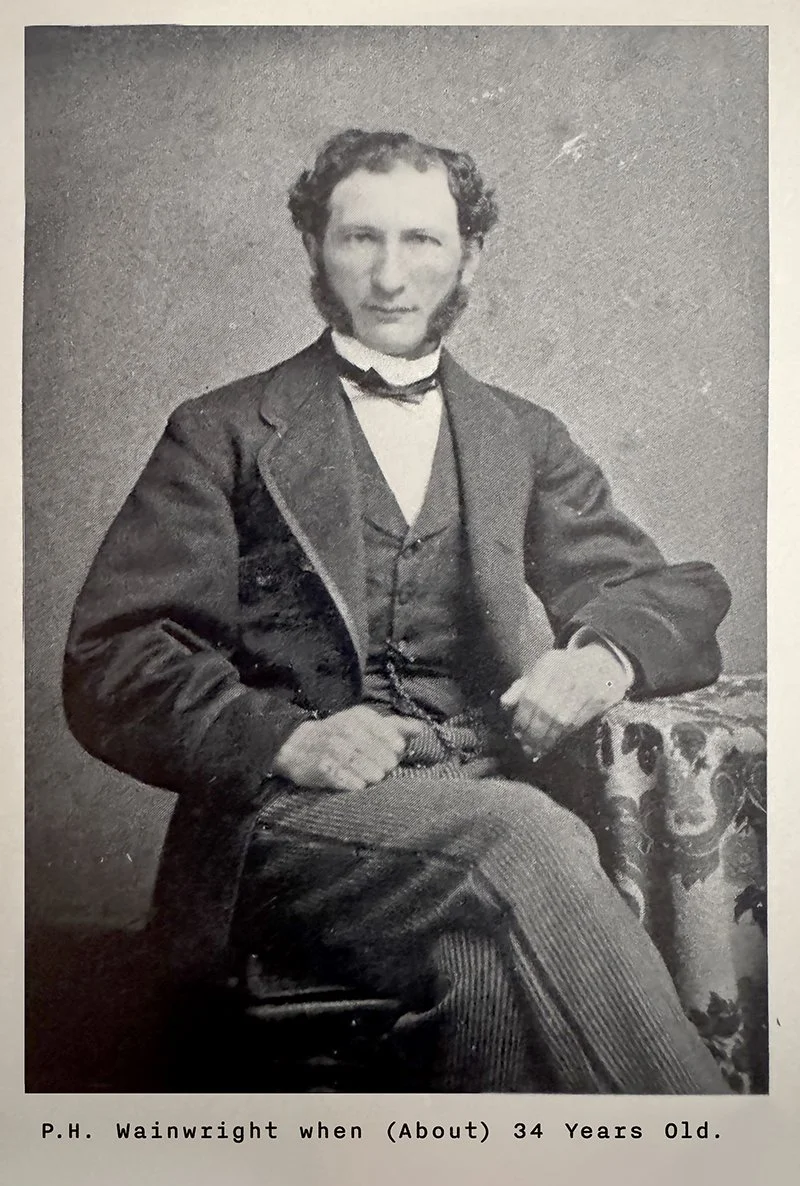 Portrait photograph of P. H. Wainwright at about age 34, seated in a dark three-piece suit with a bow tie, sporting prominent sideburns and resting one arm on a patterned upholstered chair.