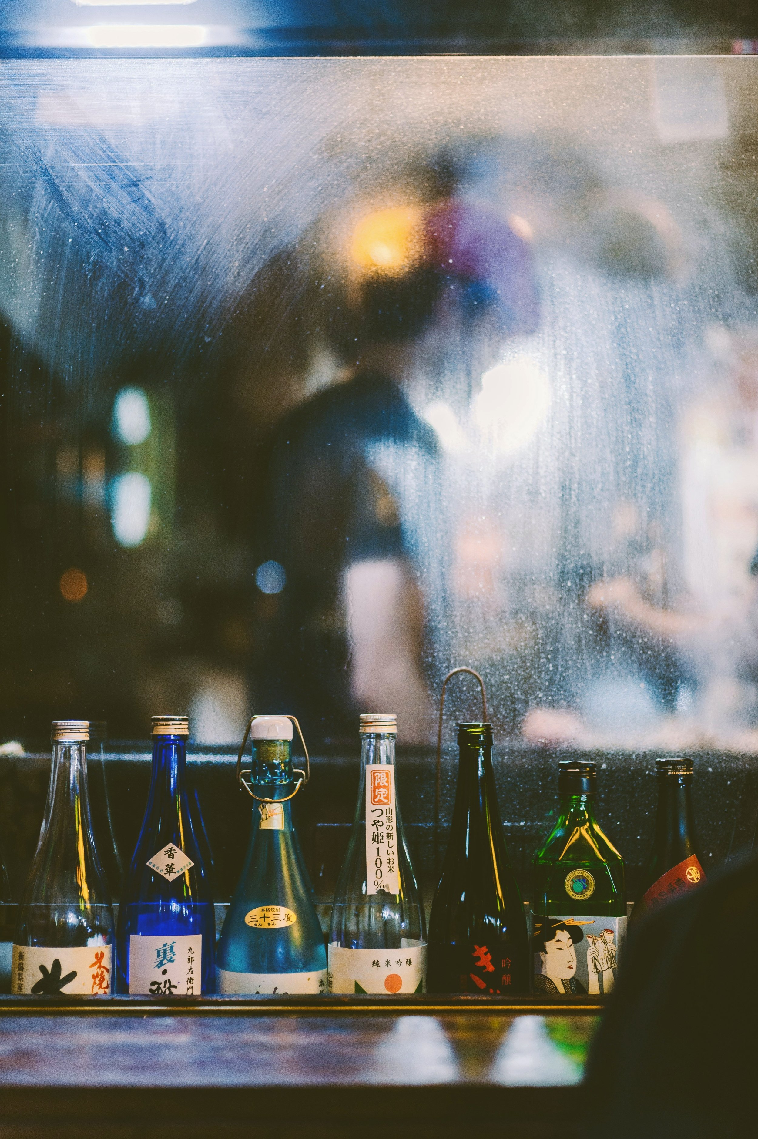 Sake bottles lined up on a shelf at an izakaya, blurred with city lights at night, Tokyo.