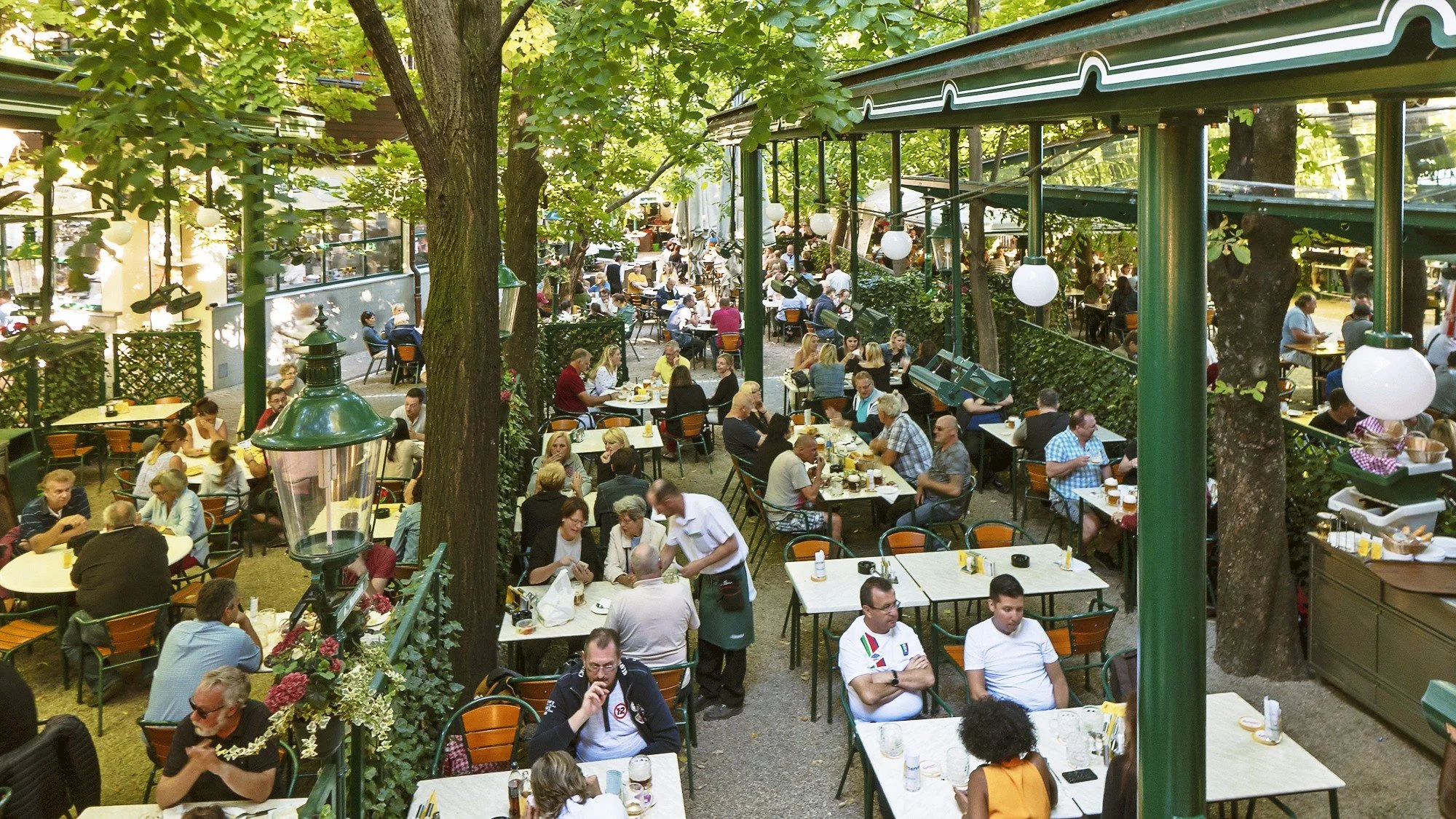 Crowded outdoor Schweizerhaus restaurant patio filled with people dining at tables surrounded by trees.