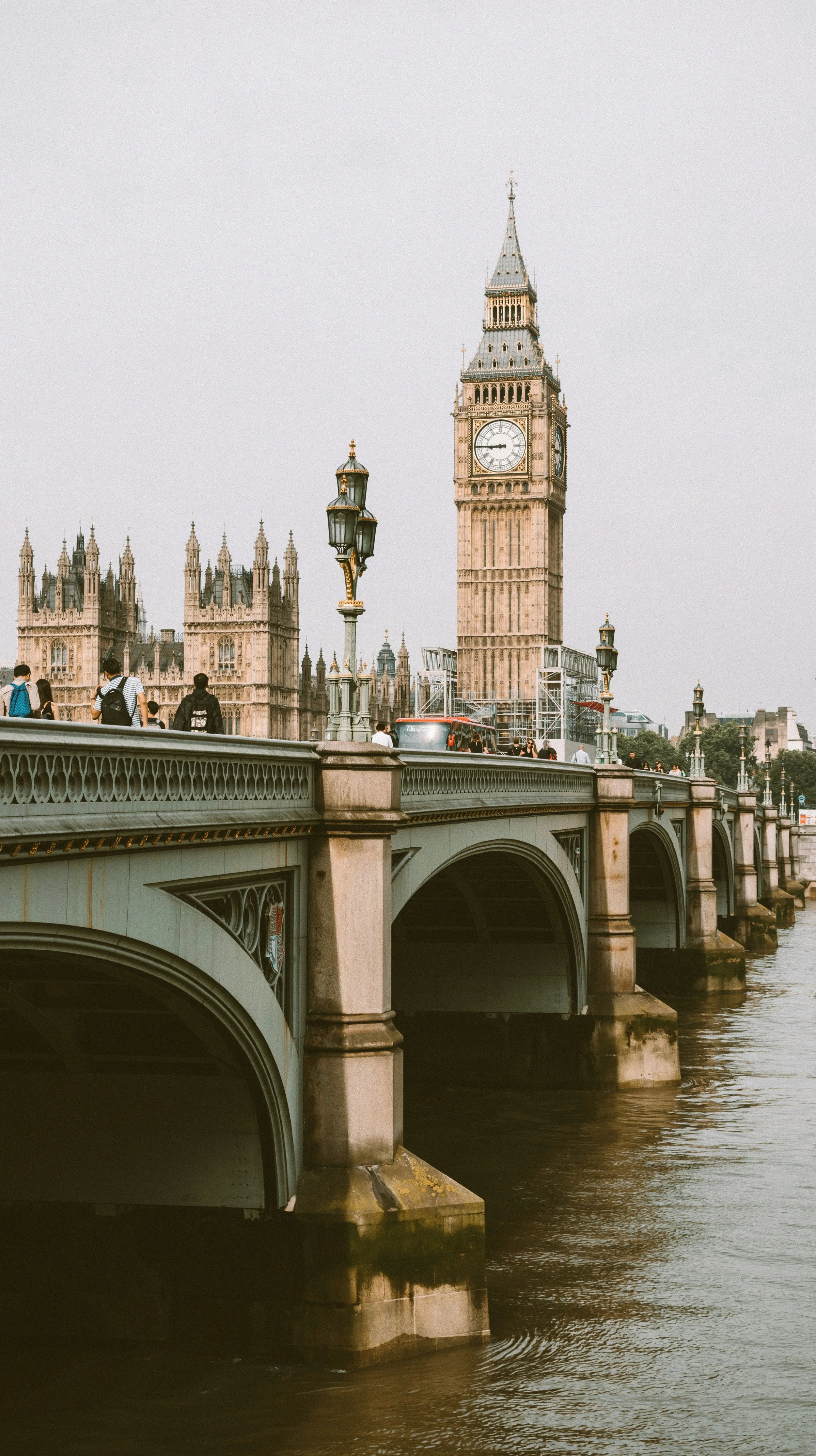 Big Ben clock tower and Westminster Palace viewed from Westminster Bridge in London, England, with pedestrians walking on the bridge and river in the foreground.