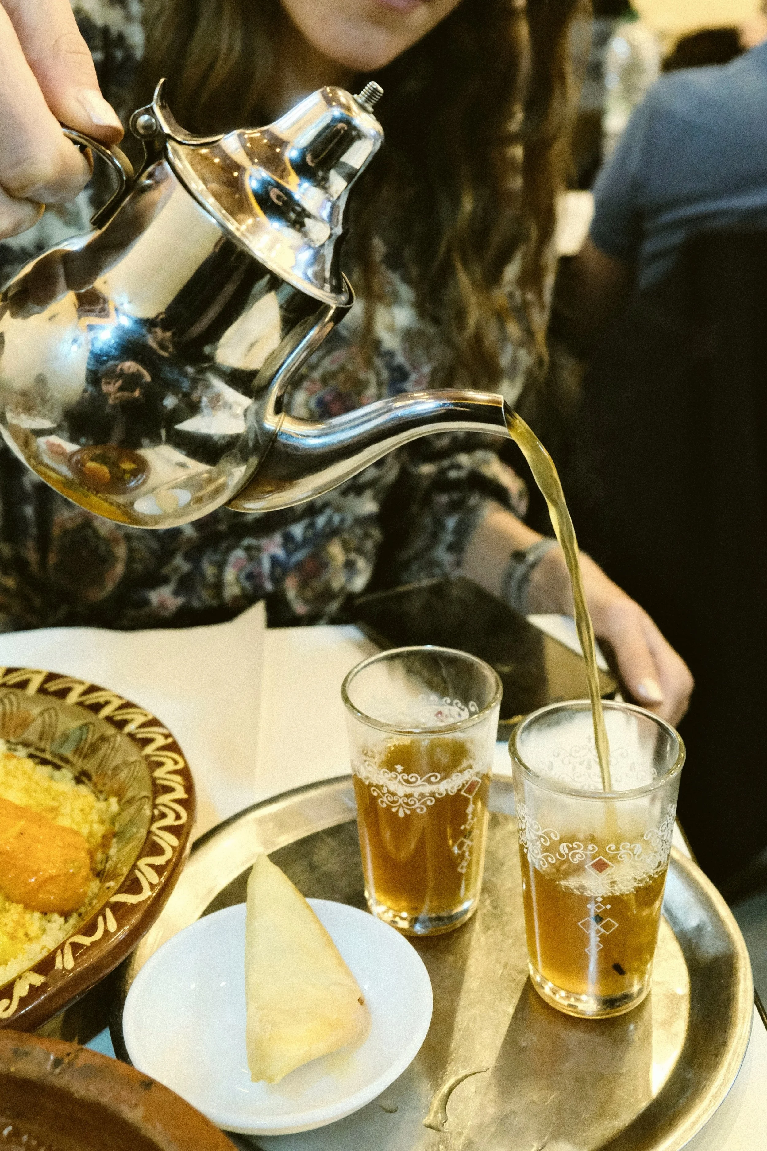 Mint tea being poured from a silver teapot into glasses beside a small plate of sweets, Marrakech.