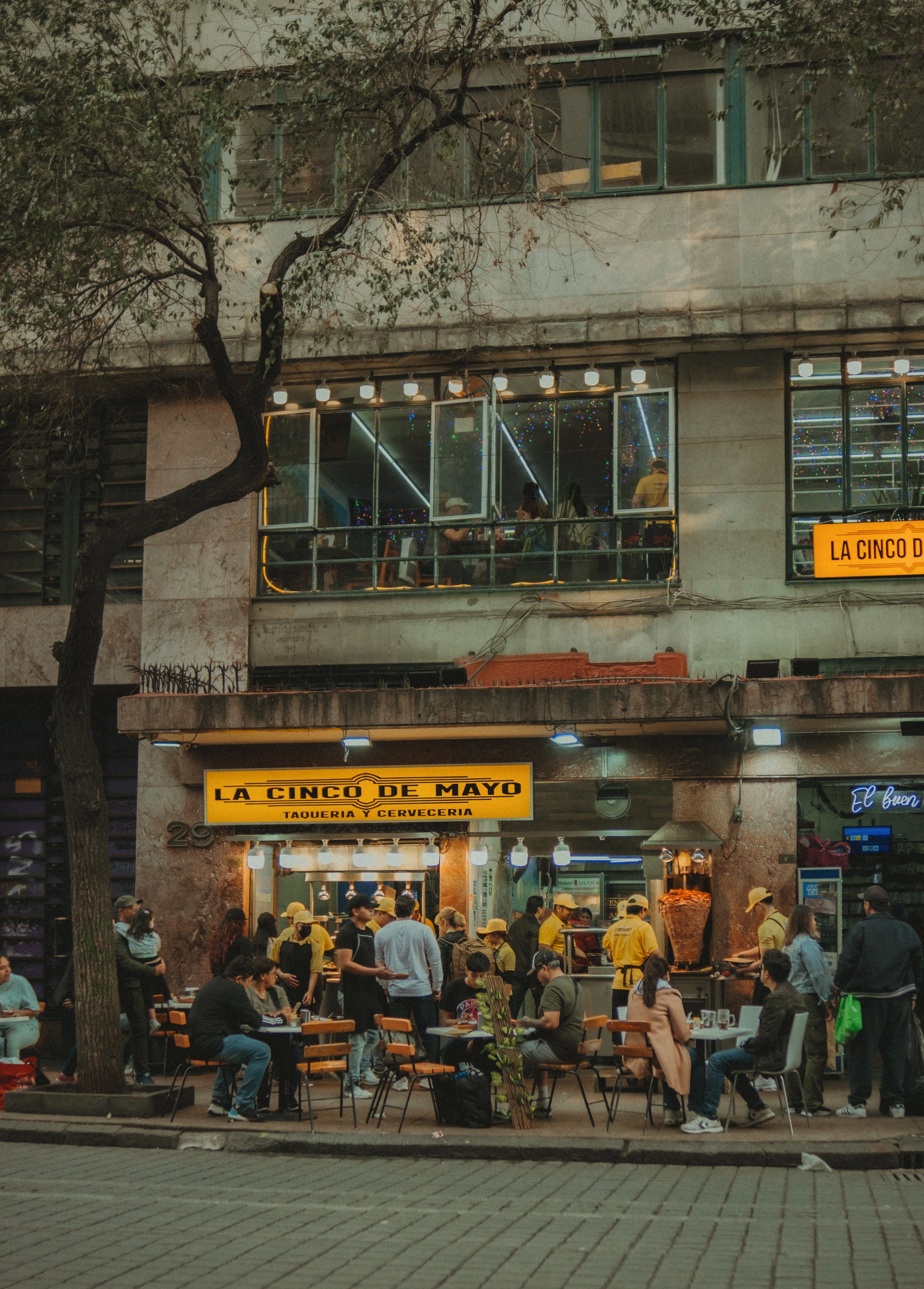 Street corner with a colorful storefront and people gathered outside, beneath a bare tree and old buildings, Mexico City.