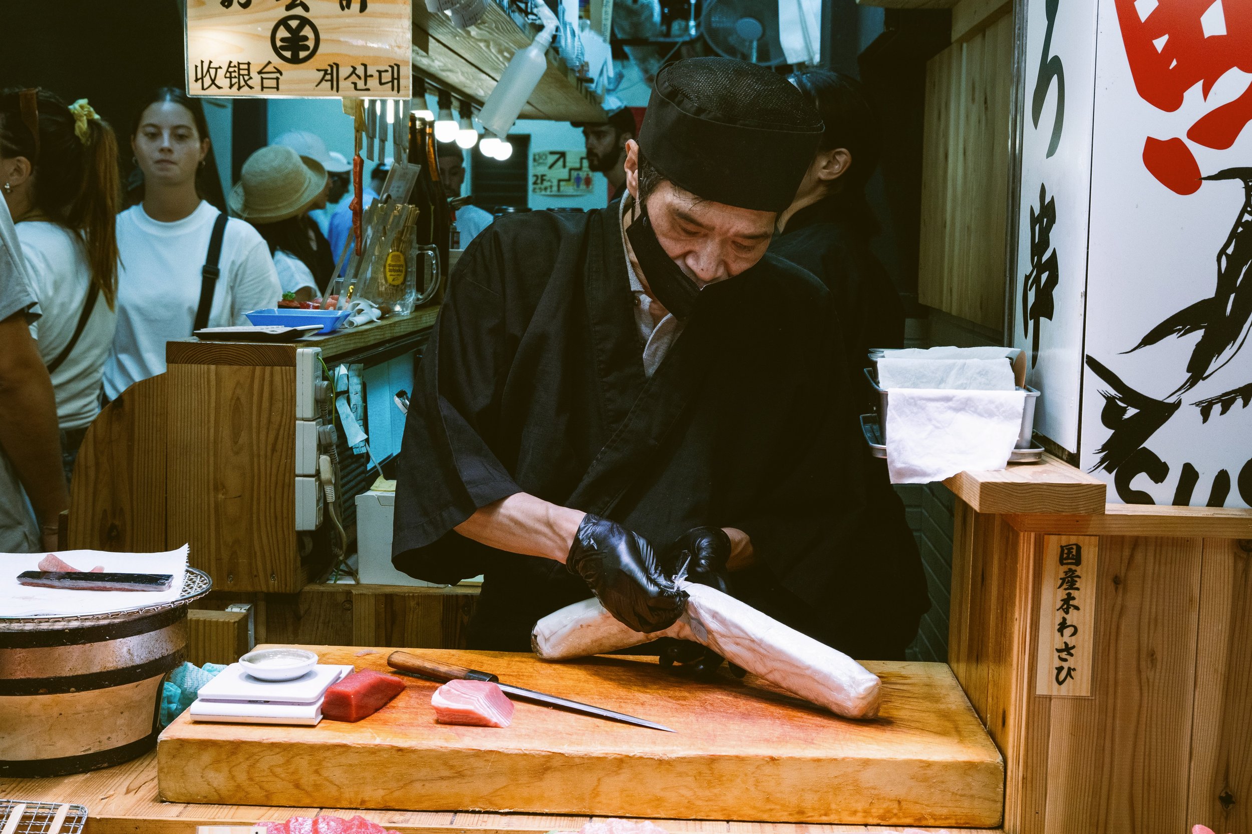 Chef in a dark uniform slicing a large fish at a market counter with onlookers behind, Tokyo.