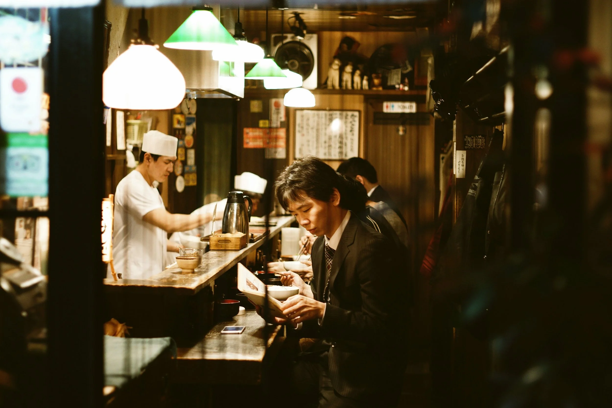 Diners seated at a wooden counter in an izakaya with a cook working behind it under warm pendant lights, Tokyo.