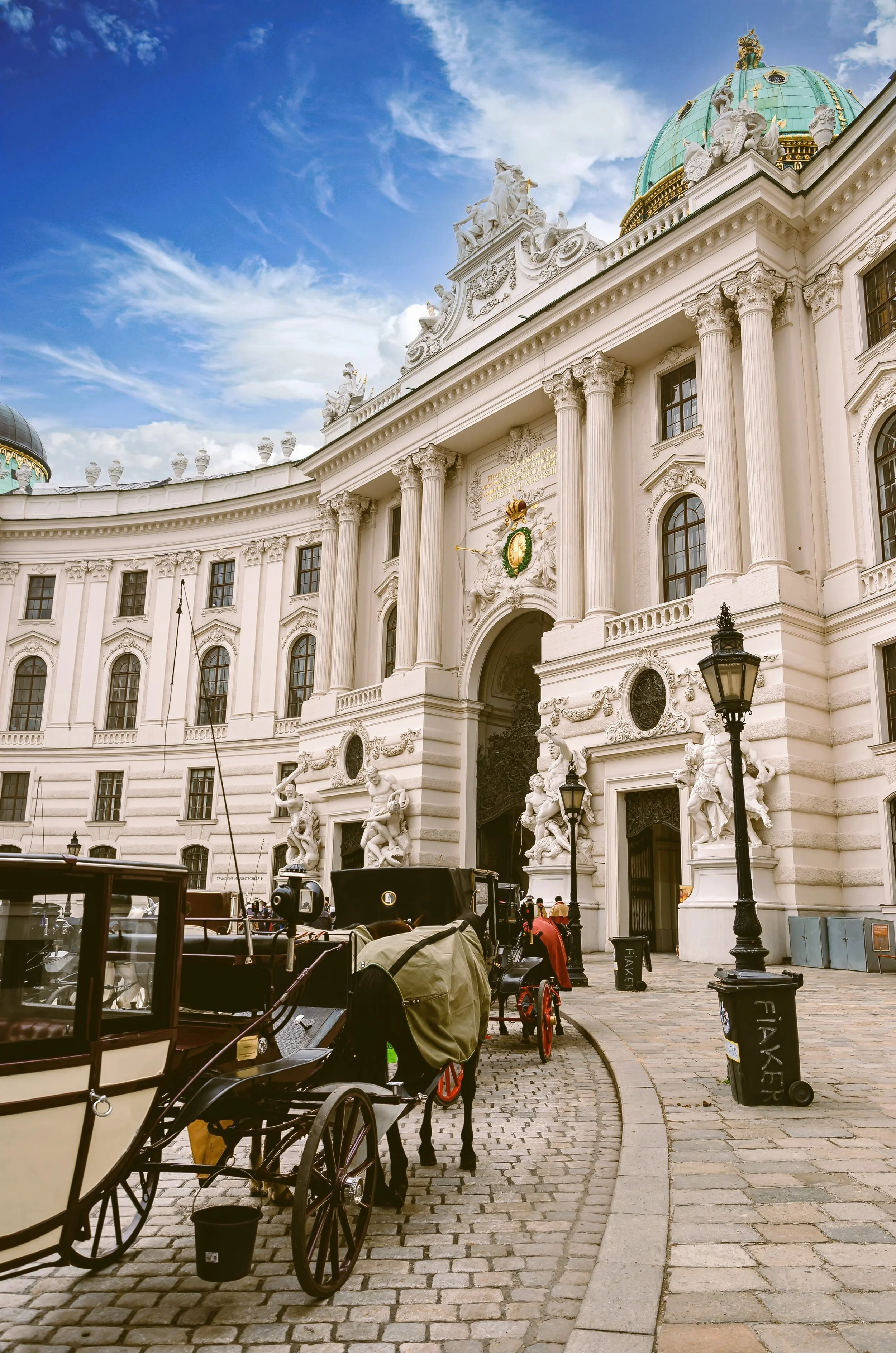 Hofburg Palace facade on Michaelerplatz with a horse-drawn carriage and fiaker in the foreground under a blue sky, Vienna.