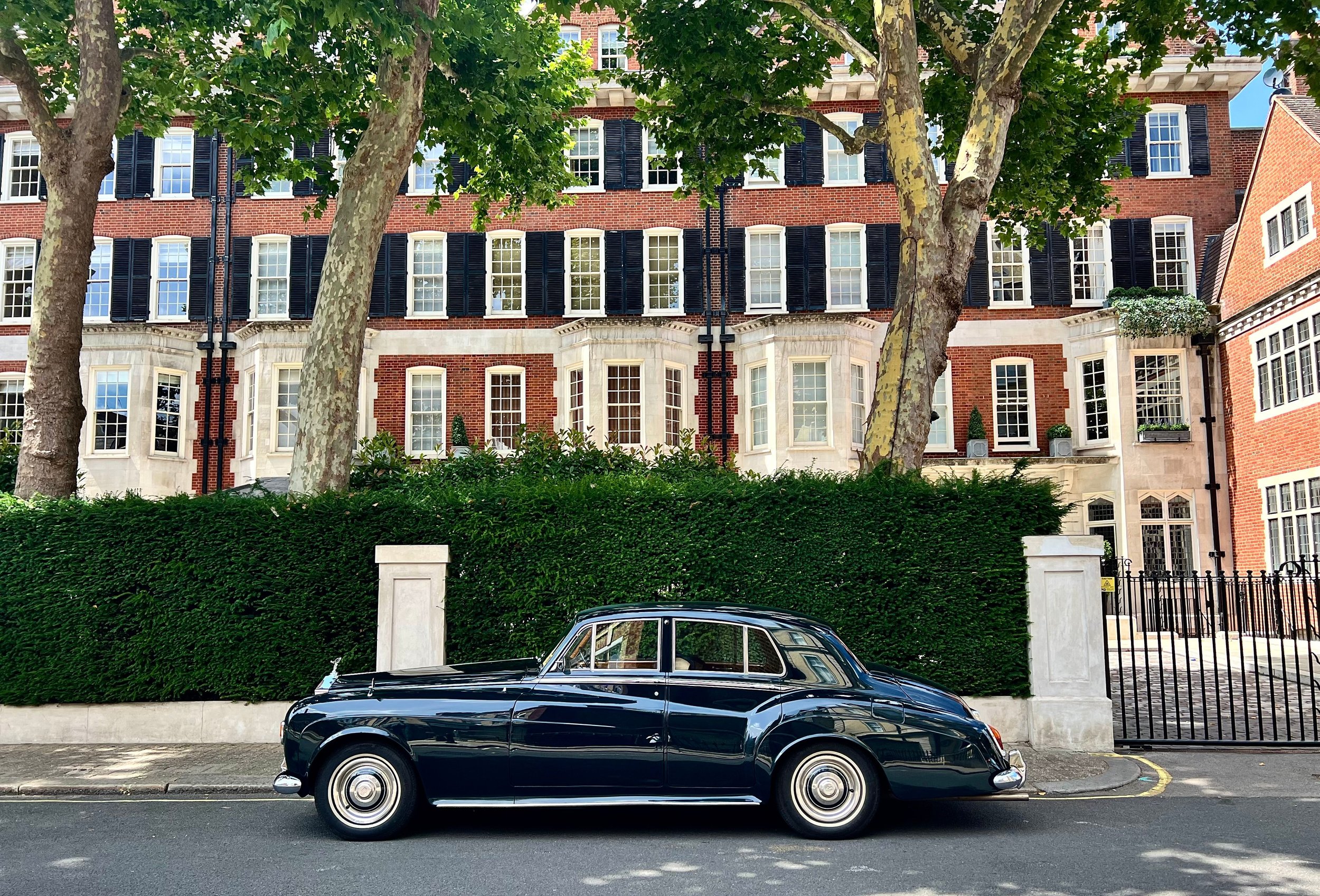 Vintage dark blue car parked in front of a red-brick terraced house behind a hedge, Chelsea, London.