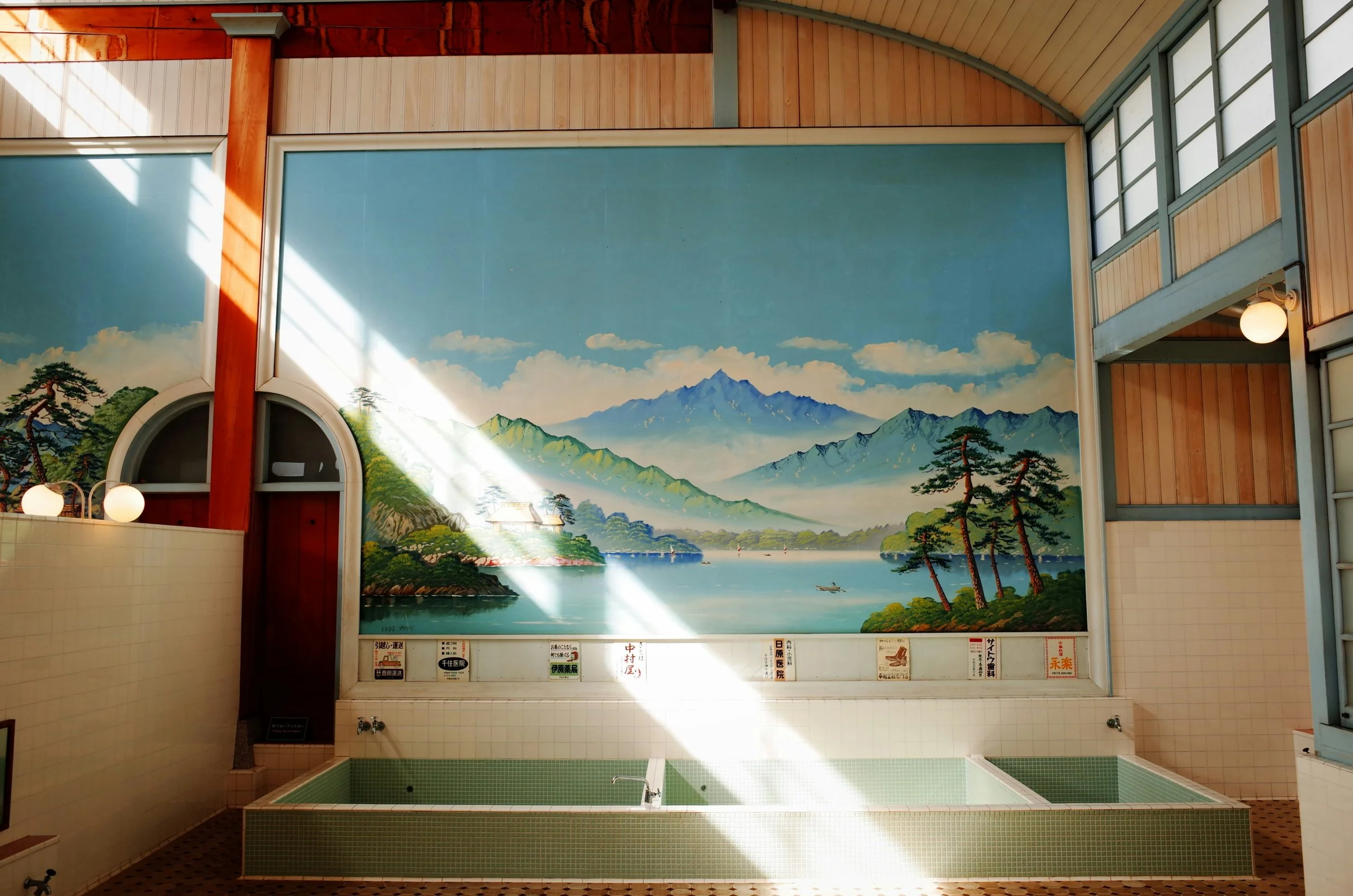Onsen bathhouse with a large painted mountain mural on the wall and sunlight streaming across the floor, Japan.