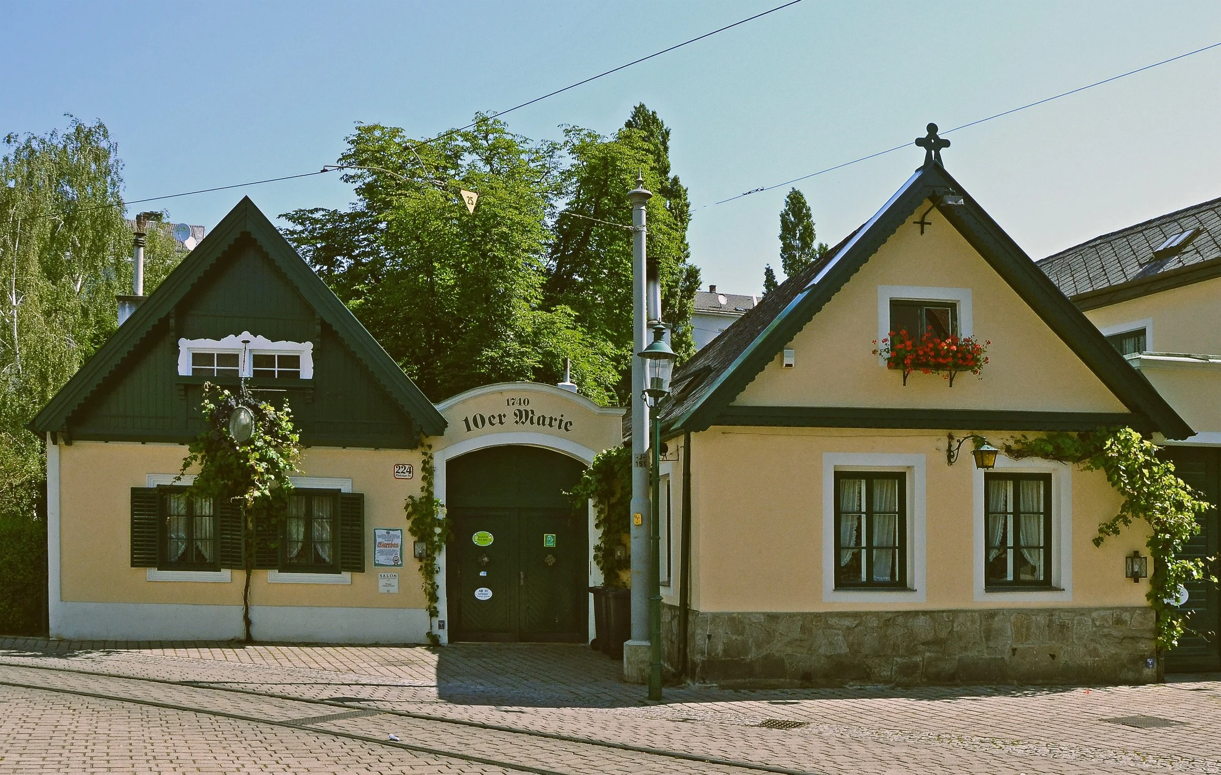A yellow house with dark shutters and a dark front door, decorated with plants and flowers. The house has steep gable roofs and Bavarian-style architectural details, with a sign reading "10er Marie" above the gate. There are trees and a streetlamp in the background, along with a clear blue sky.