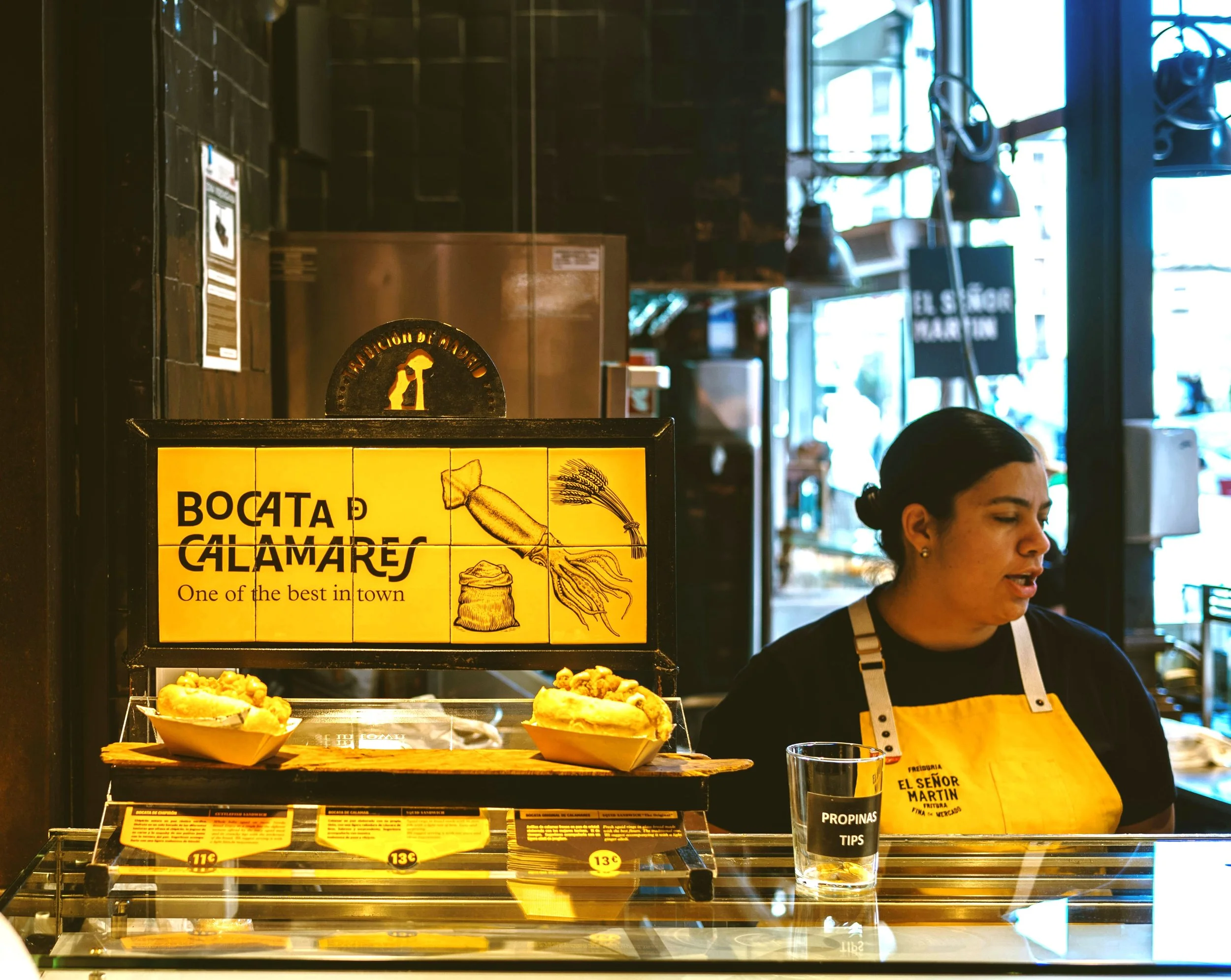 Calamari sandwiches on a counter beneath a yellow tiled "Bocata de Calamares" sign, Madrid