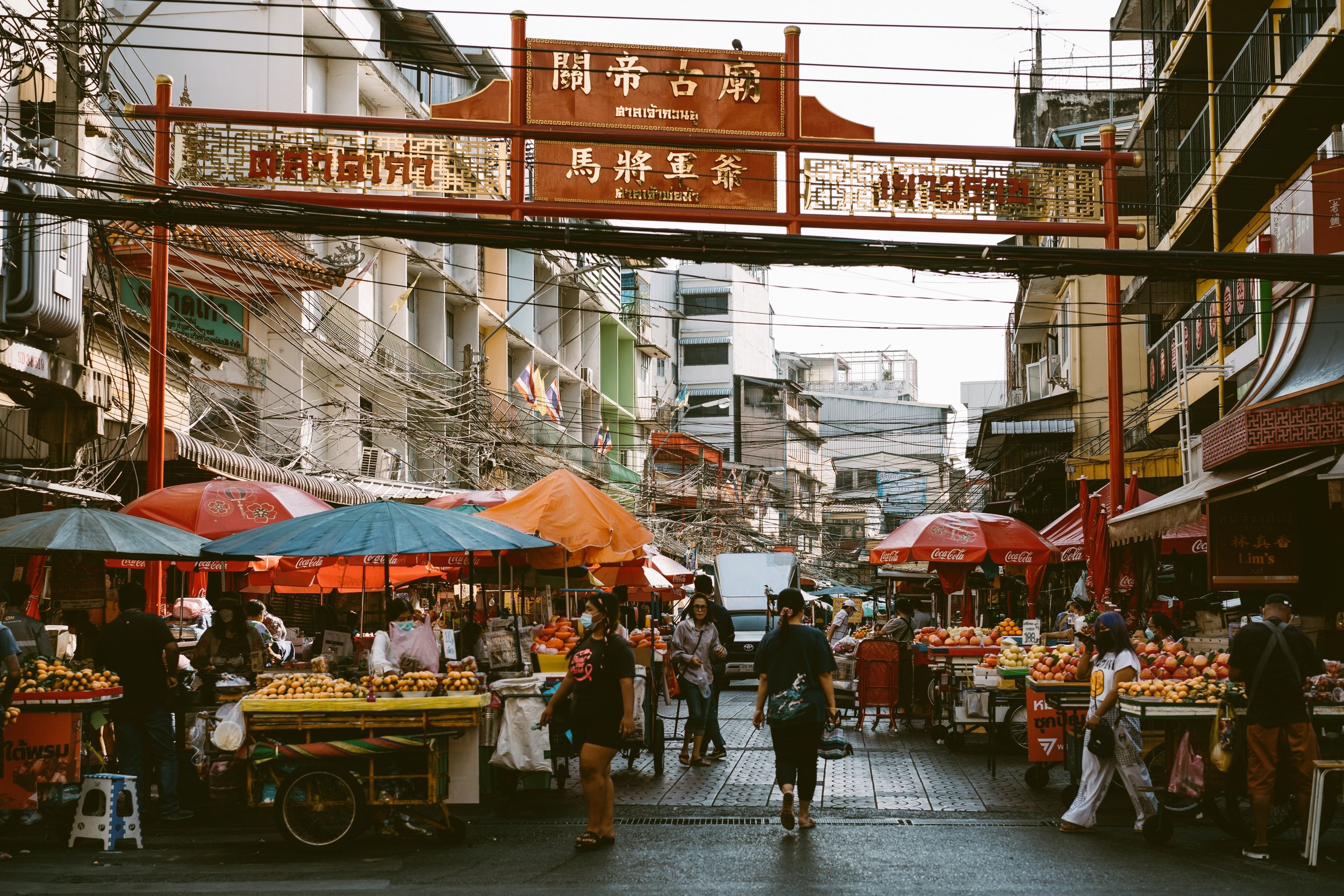 Street market under colorful umbrellas with a red Chinese-style gate above the road, Chinatown, Bangkok.