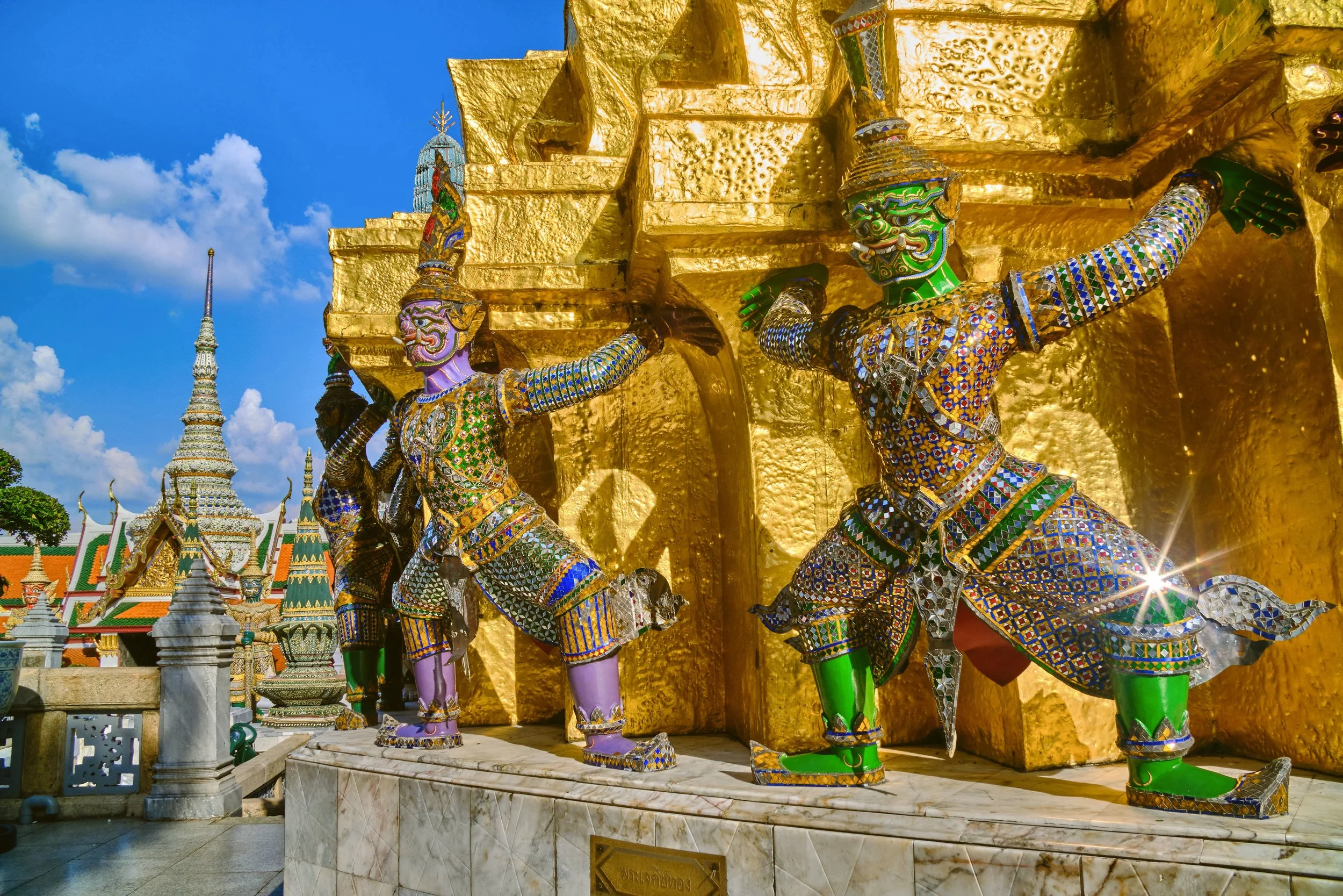 Colorful demon guardian statues holding up a golden chedi under a blue sky, Grand Palace, Bangkok.