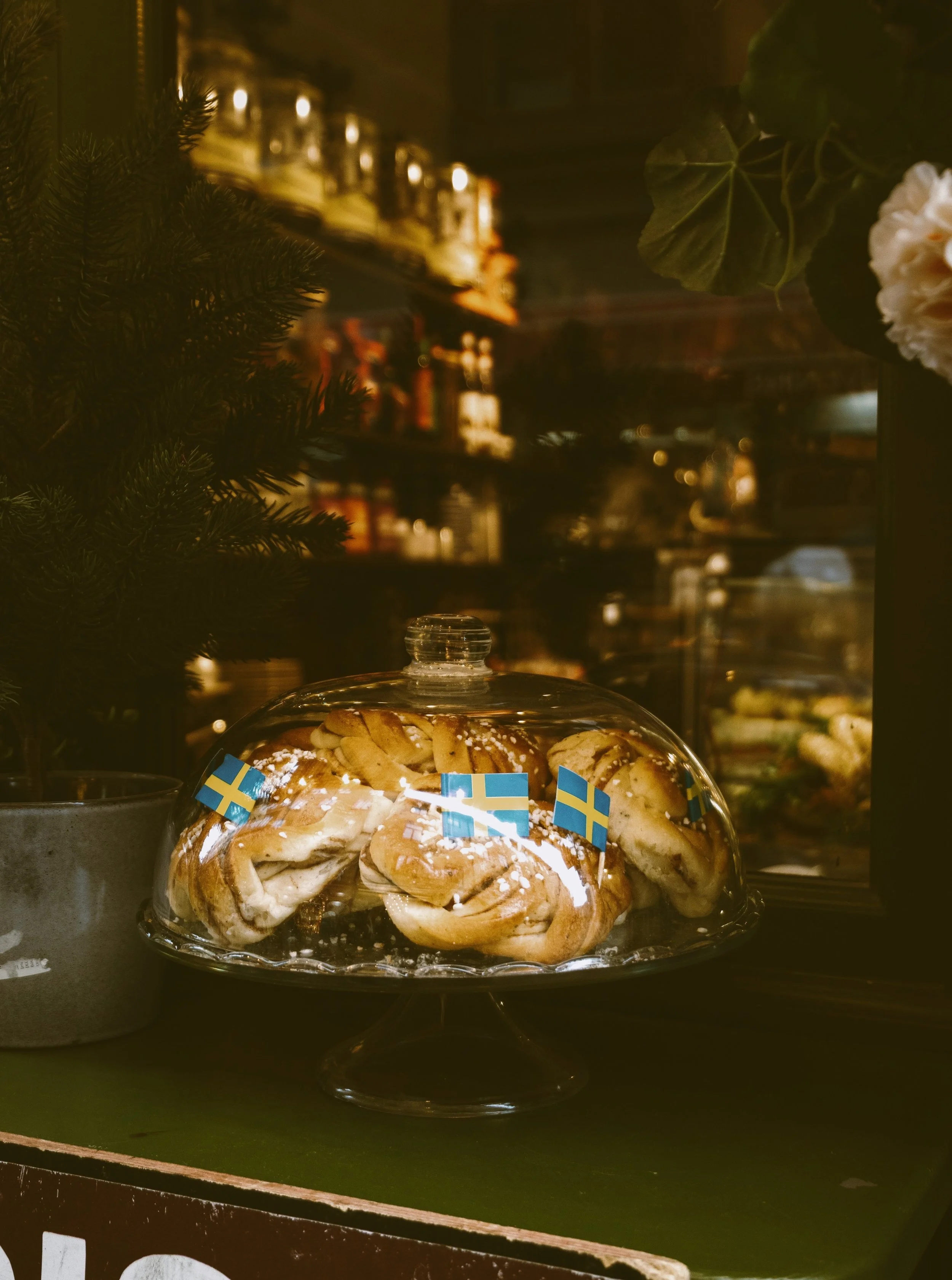 A glass cake stand with cinnamon rolls decorated with Swedish flags inside a bakery or café.