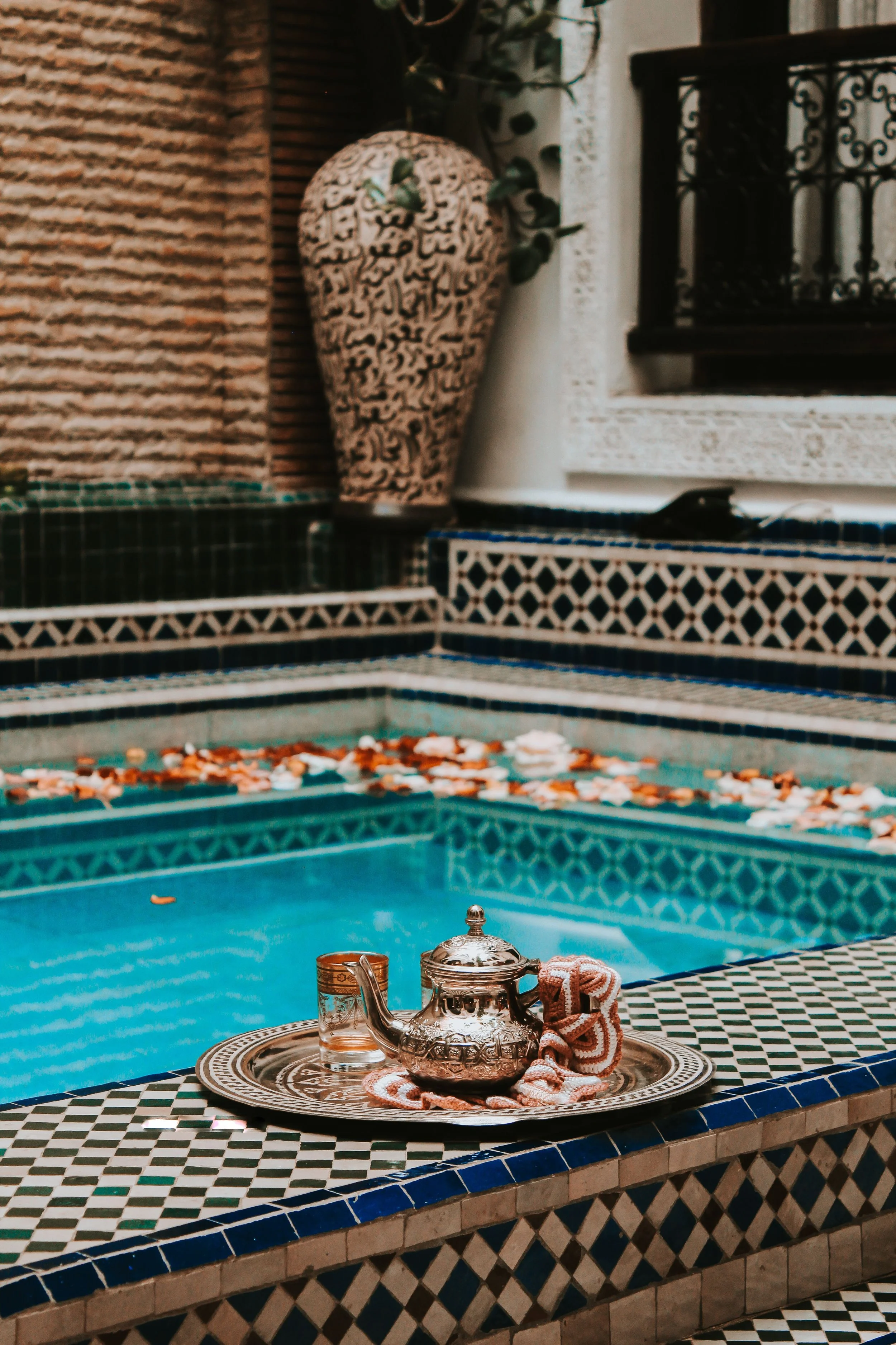 Turquoise pool with a decorated tray on its edge and ornate tilework beside a clay urn, Marrakech.