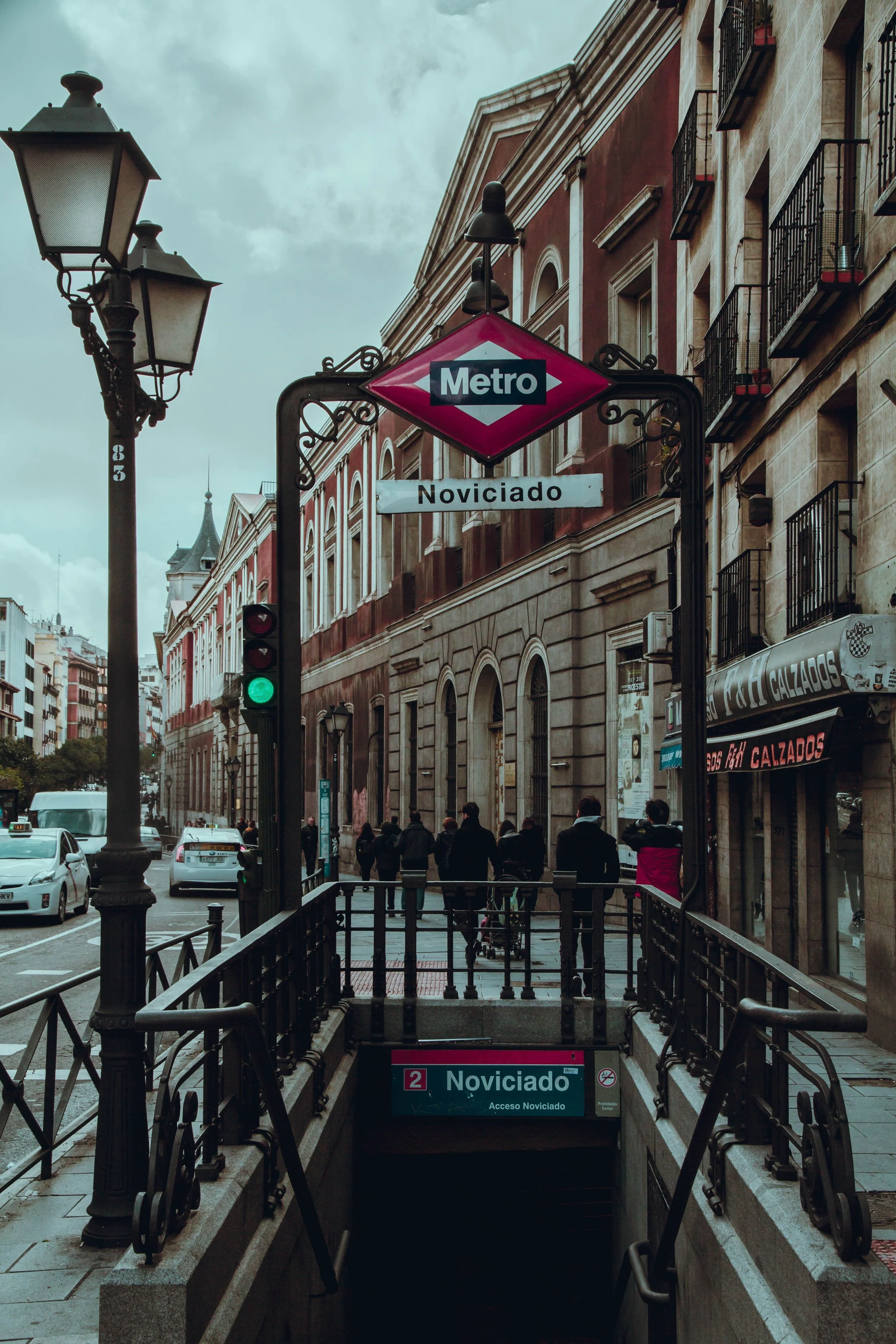 Entrance to Noviciado metro station with a street scene, cars, buildings, and people walking.
