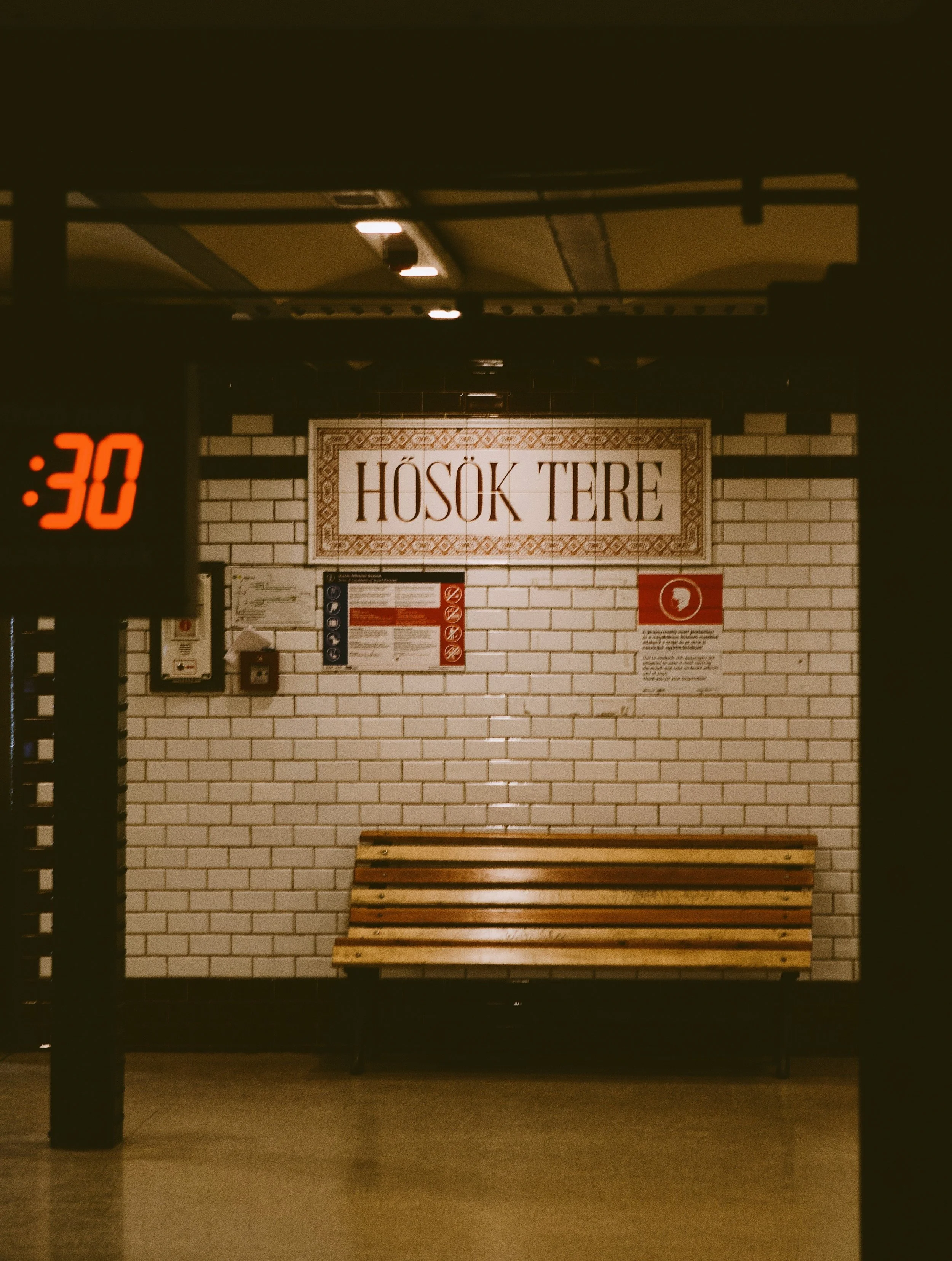 Image of a subway or train station platform with a bench, brick wall, a sign that reads 'HŌSÖK TÉRE', and an electronic display showing ':30' indicating the time.