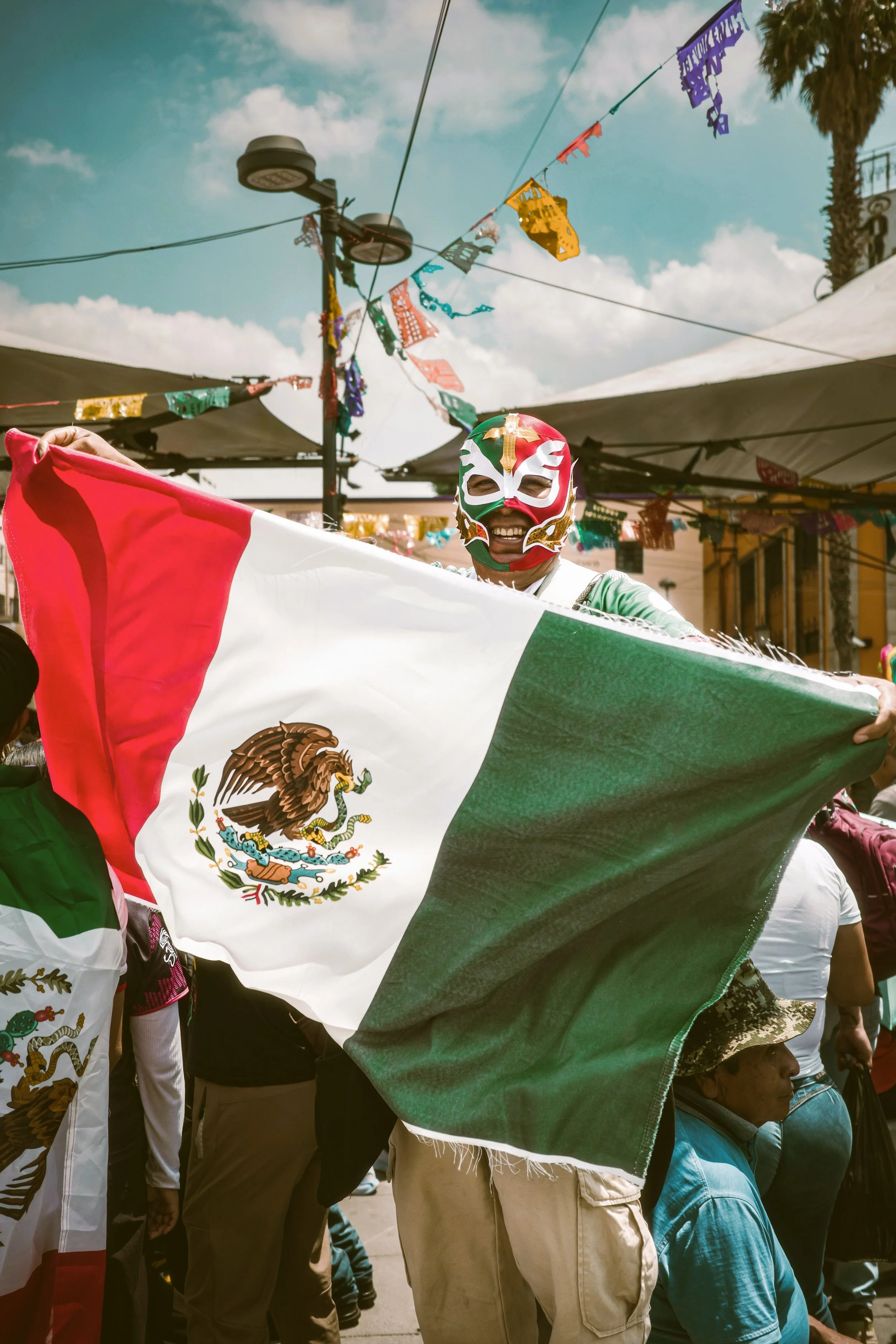 People participating in a lucha libre celebration, holding a Mexican flag, with colorful papel picado flags hanging overhead, and a partly cloudy sky in the background, Mexico City.