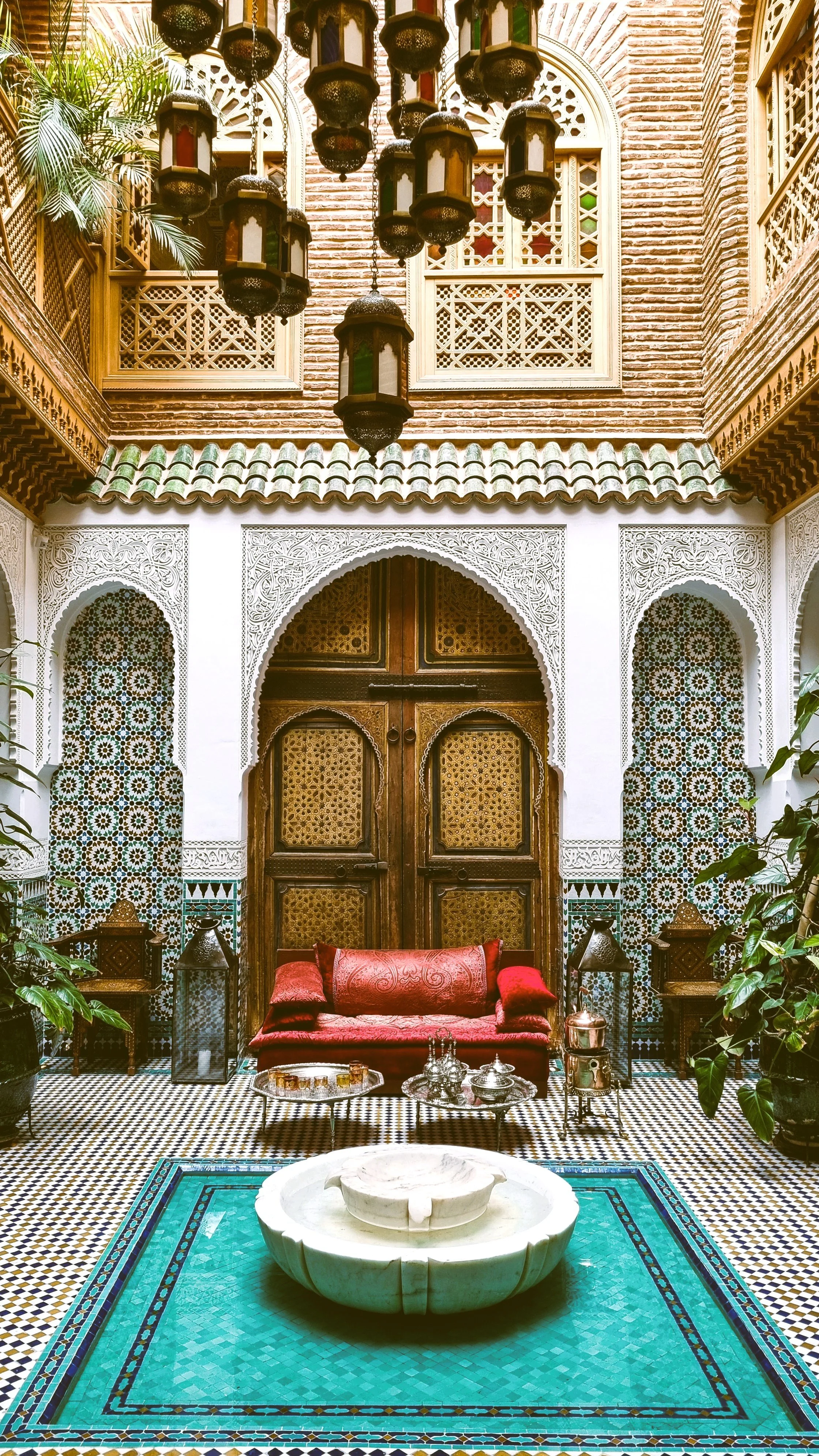 Riad courtyard with a central fountain, turquoise tiles and a red lounge area between carved arches, Marrakech.