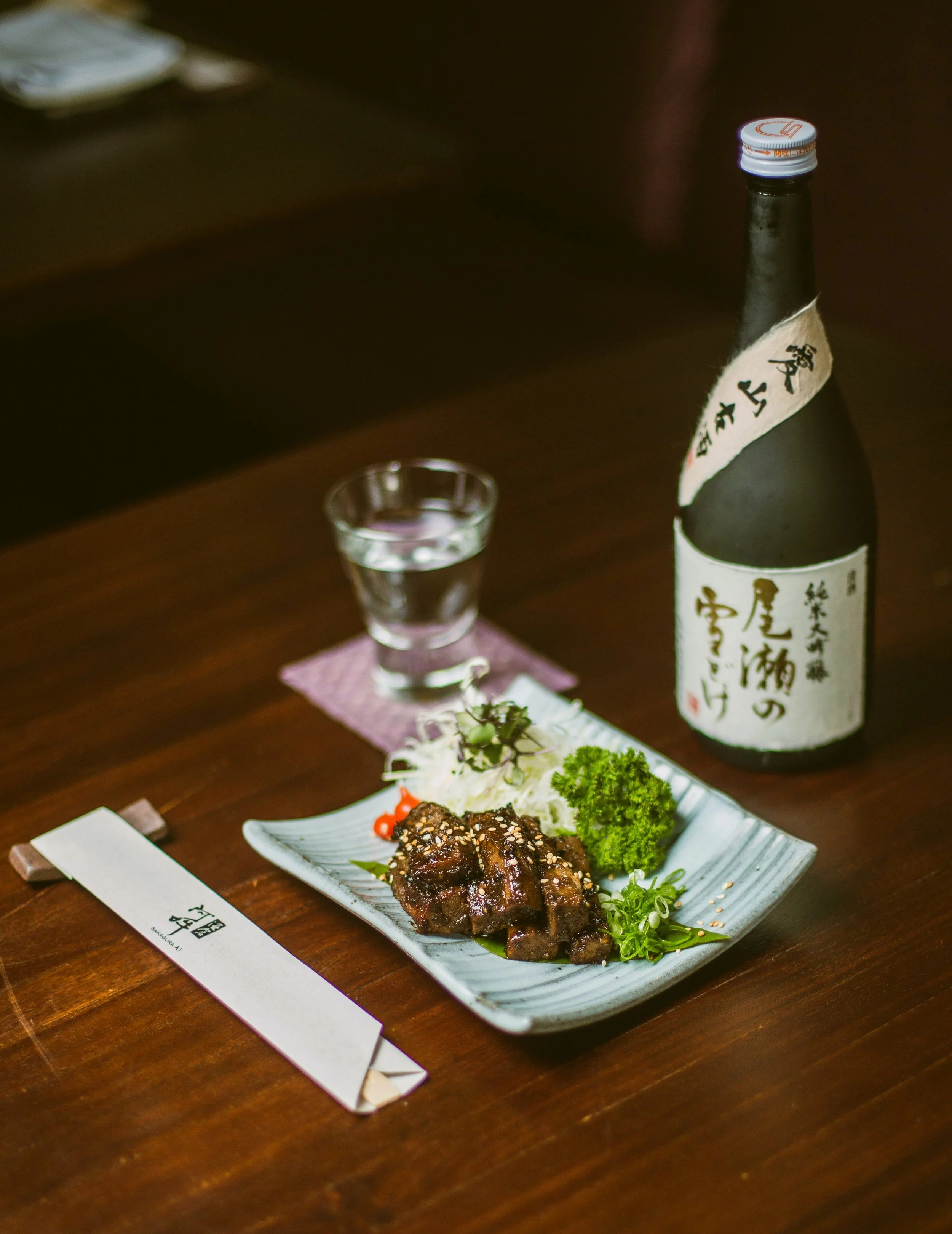 A plate of Japanese grilled meat garnished with greens, with a bottle of sake and a glass on a wooden table at a restaurant, Tokyo.