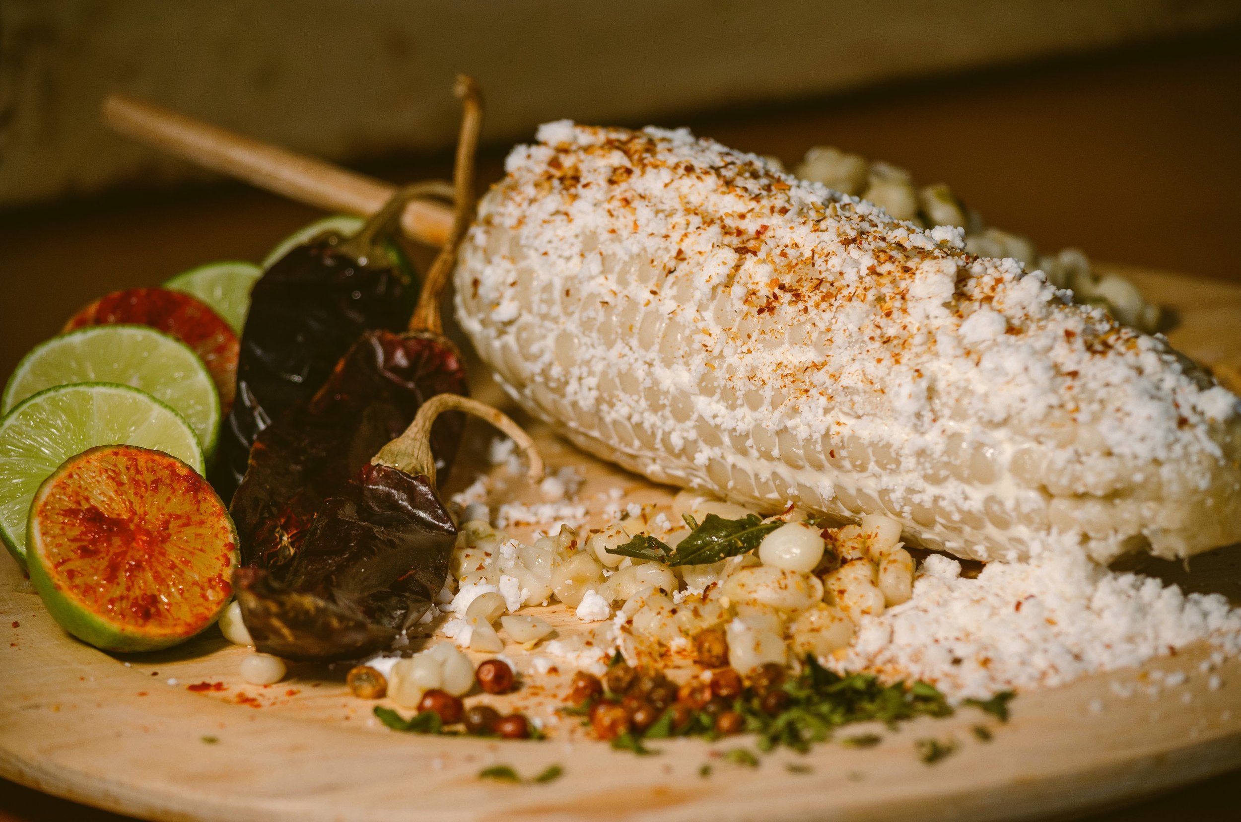 Elote covered in cheese and chili powder on a plate with grilled peppers and lime halves, Mexico City.
