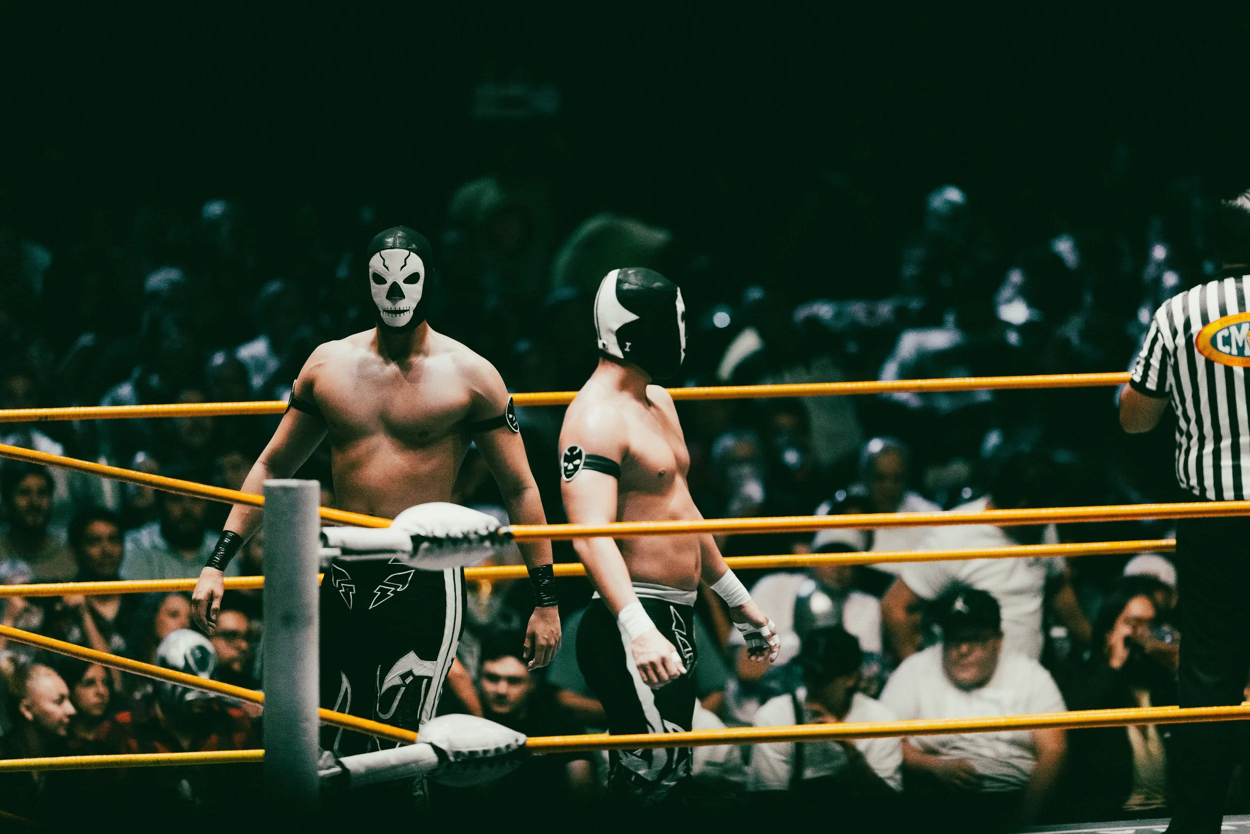 Professional lucha libre wrestlers wearing skeleton masks standing in the ring, while a referee stands nearby and a crowd watches in the background, Mexico City.