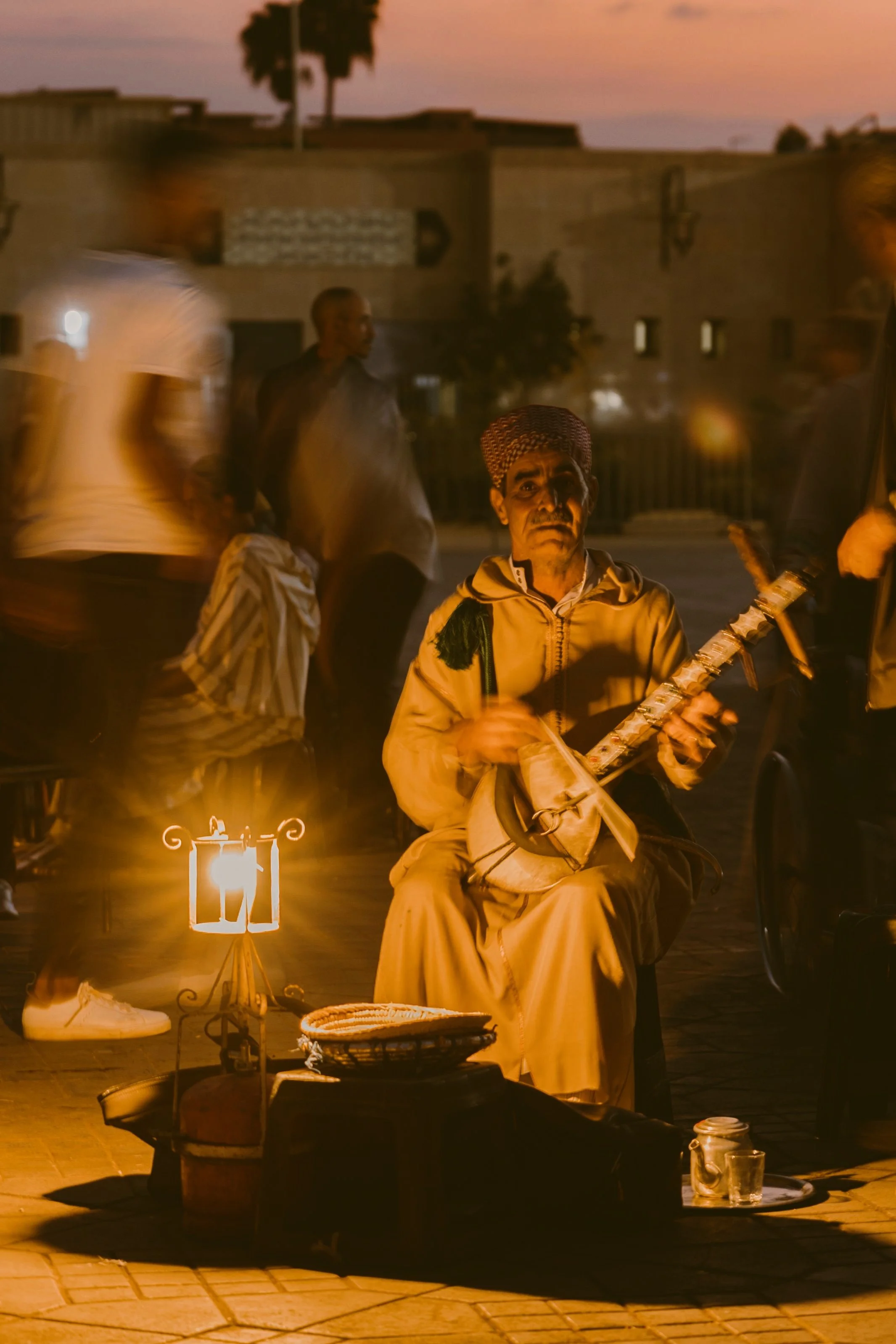 Musician playing a traditional stringed instrument beside a glowing lantern at dusk, Marrakech.