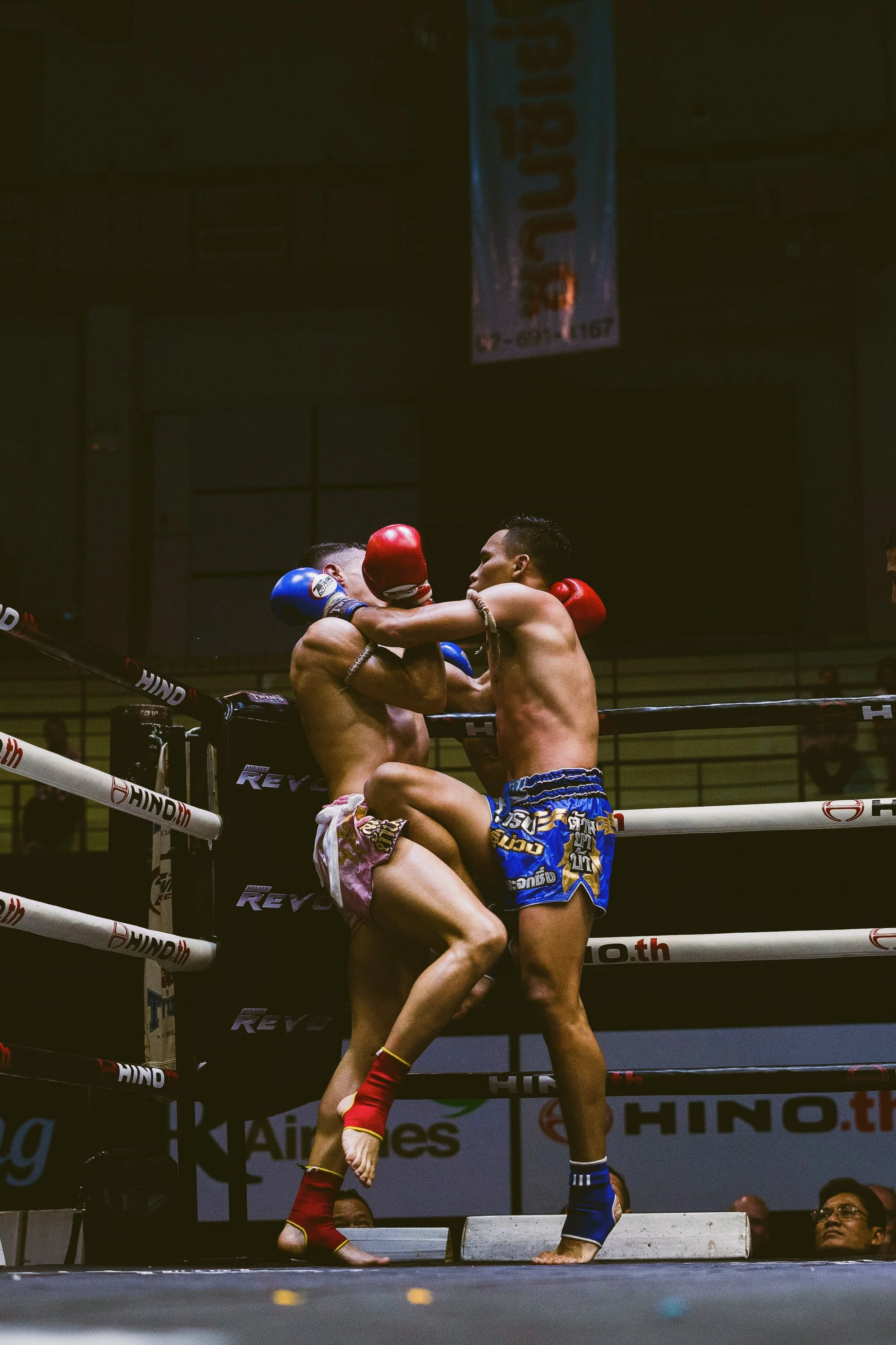 Two Muay Thai fighters in a ring engaging in a close combat exchange, surrounded by a dark background, with spectators observing.