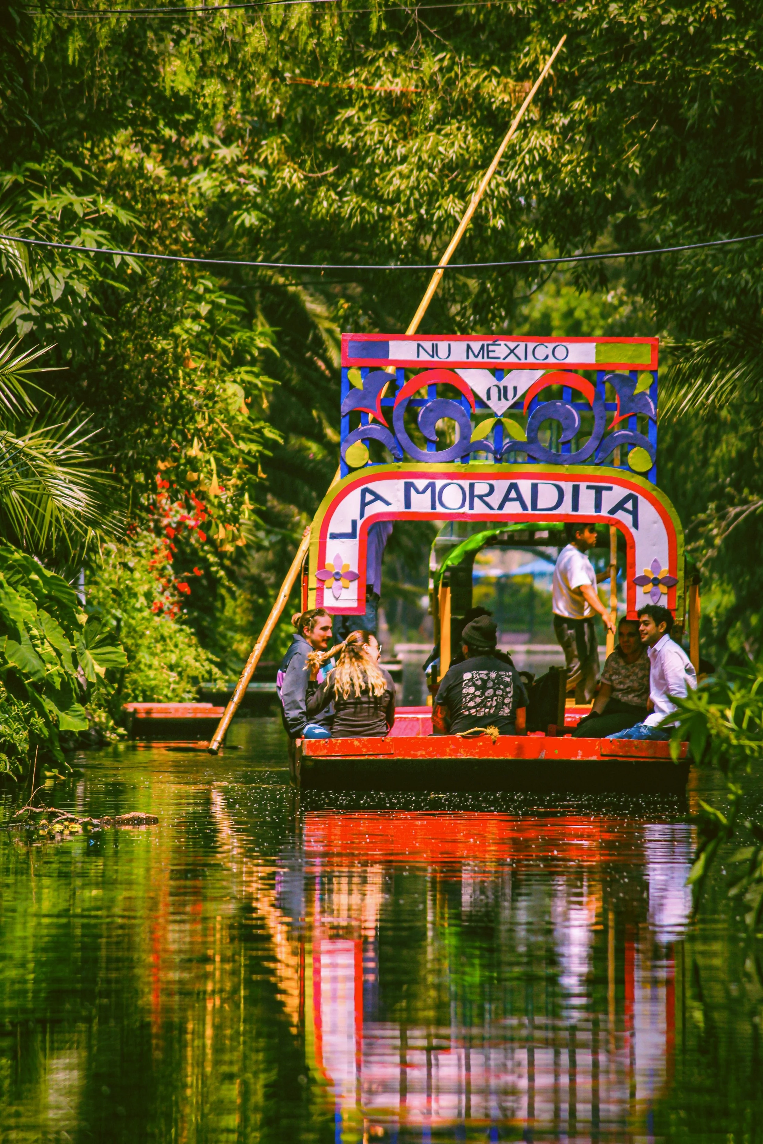 Colorful trajinera reflected in the calm water of a canal lined with lush greenery, Xochimilco, Mexico City.