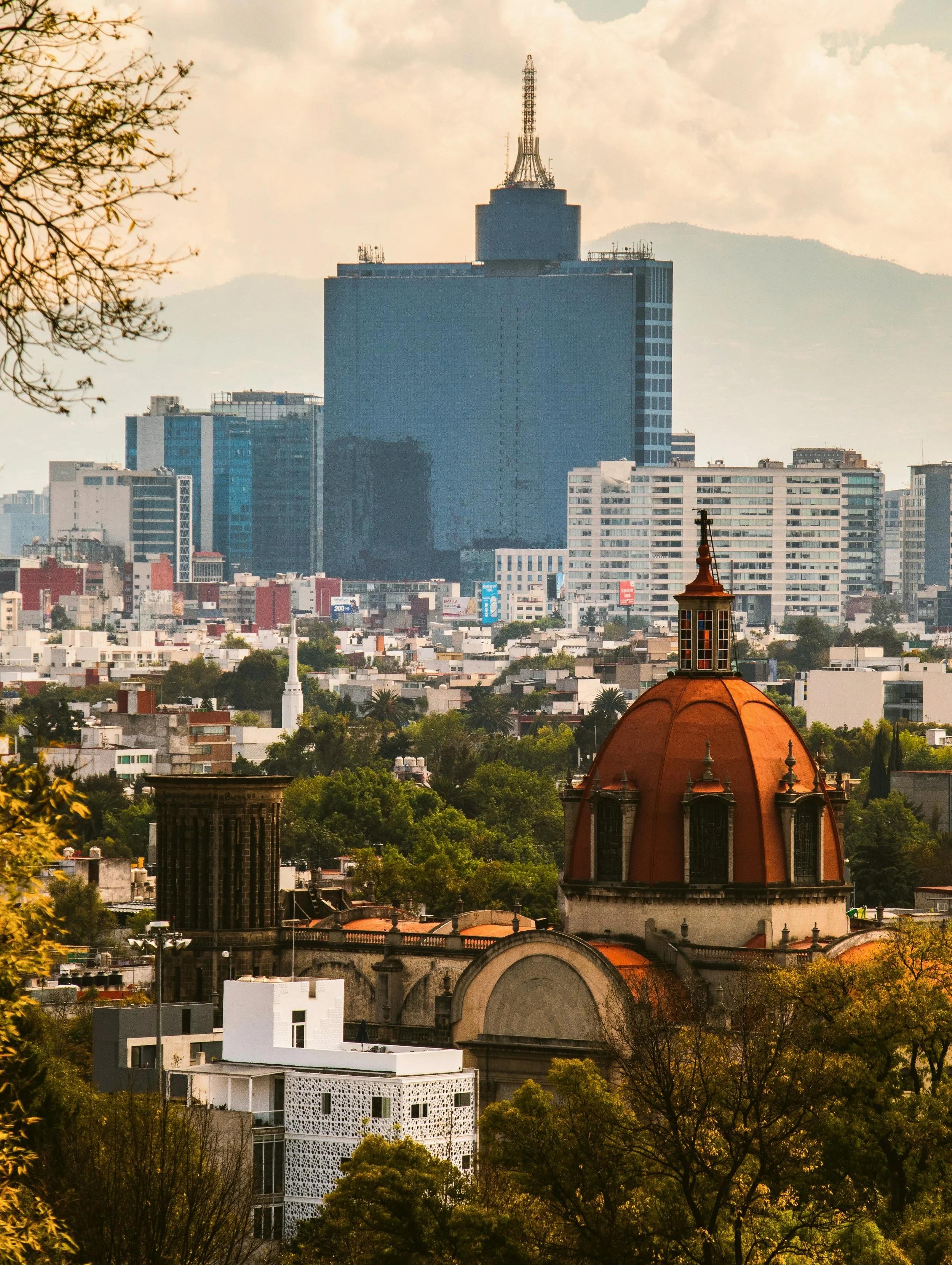 City skyline with a modern glass tower rising behind rooftops and a red-domed church, Mexico City.