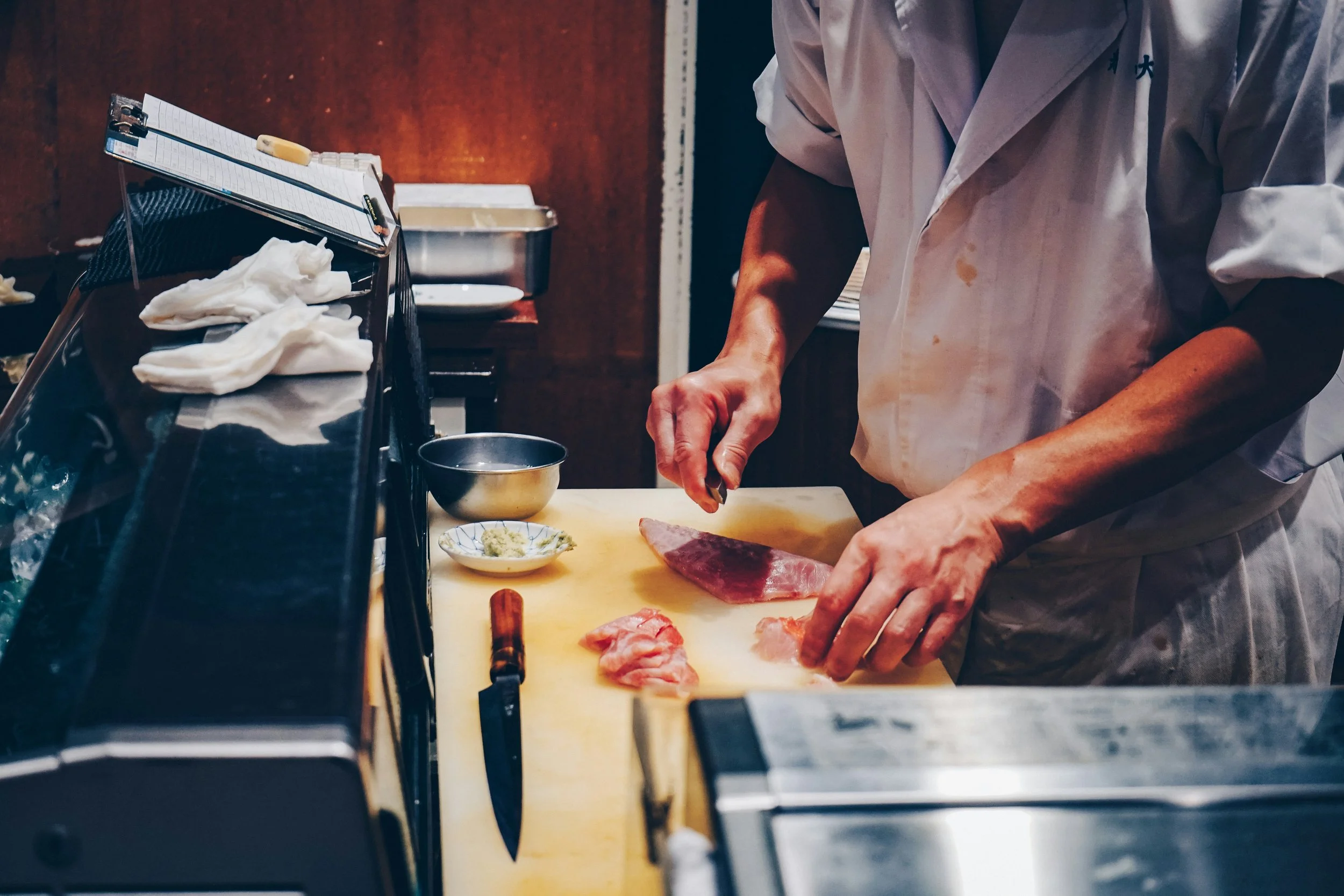 Sushi chef preparing fish at a counter with knives and equipment in the foreground, Tokyo.