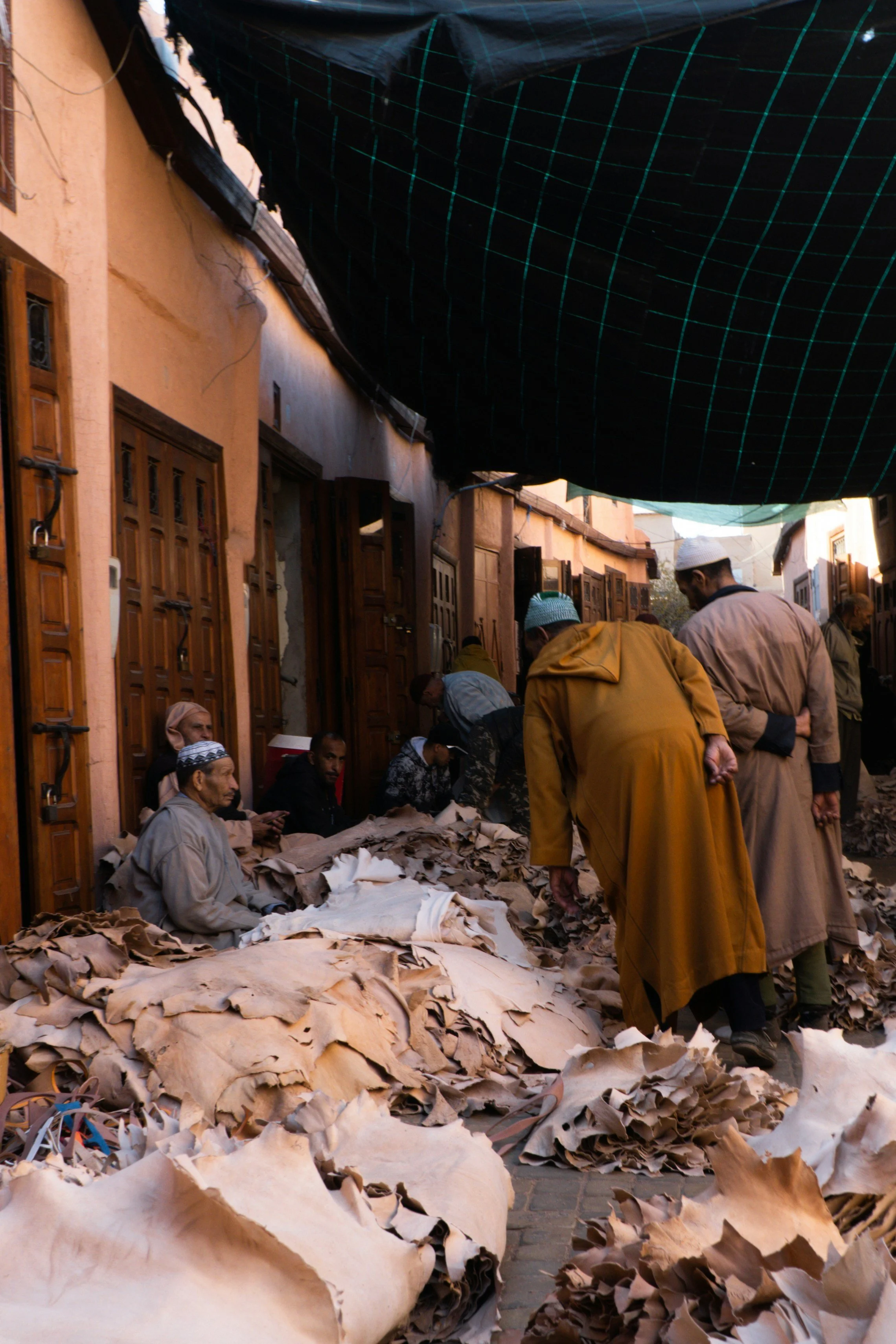 Narrow souk alley with a person in a yellow robe beside piles of goods on the ground, Marrakech.