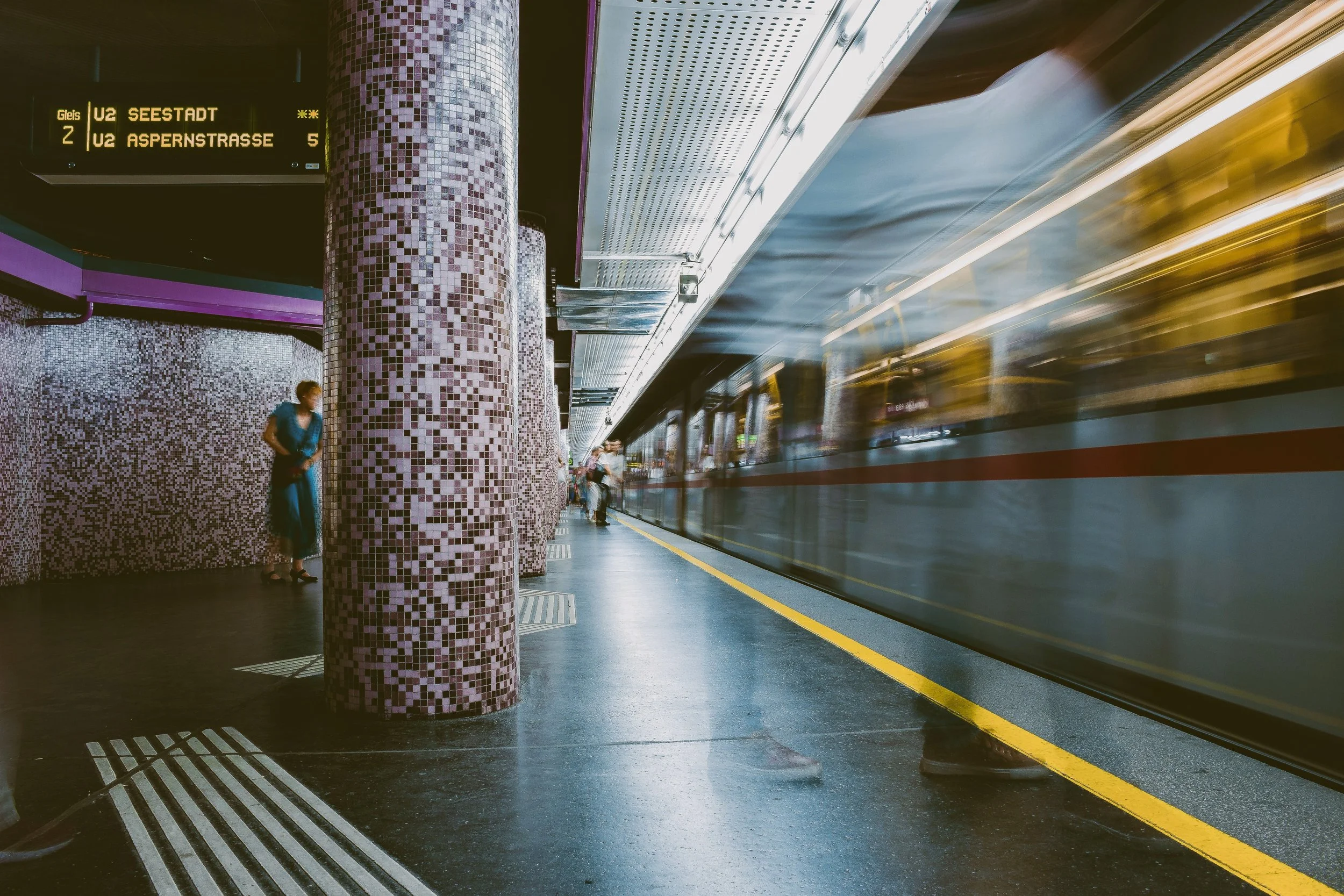 A busy Vienna metro station platform with a moving train on the right side, and passengers waiting.