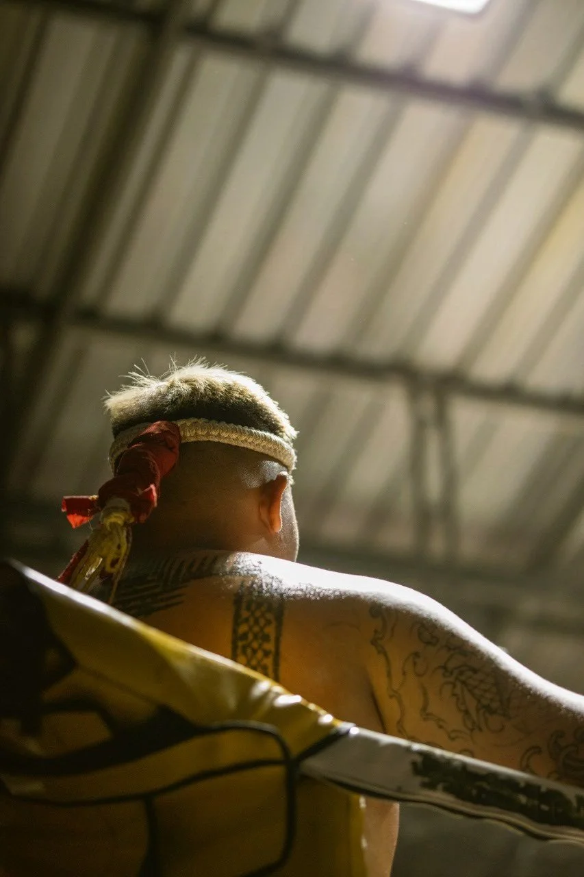Back view of a Muay Thai fighter with a traditional topknot hairstyle, tattoos on their shoulder and arm, and a red band tied around their head, inside a metal roof structure.