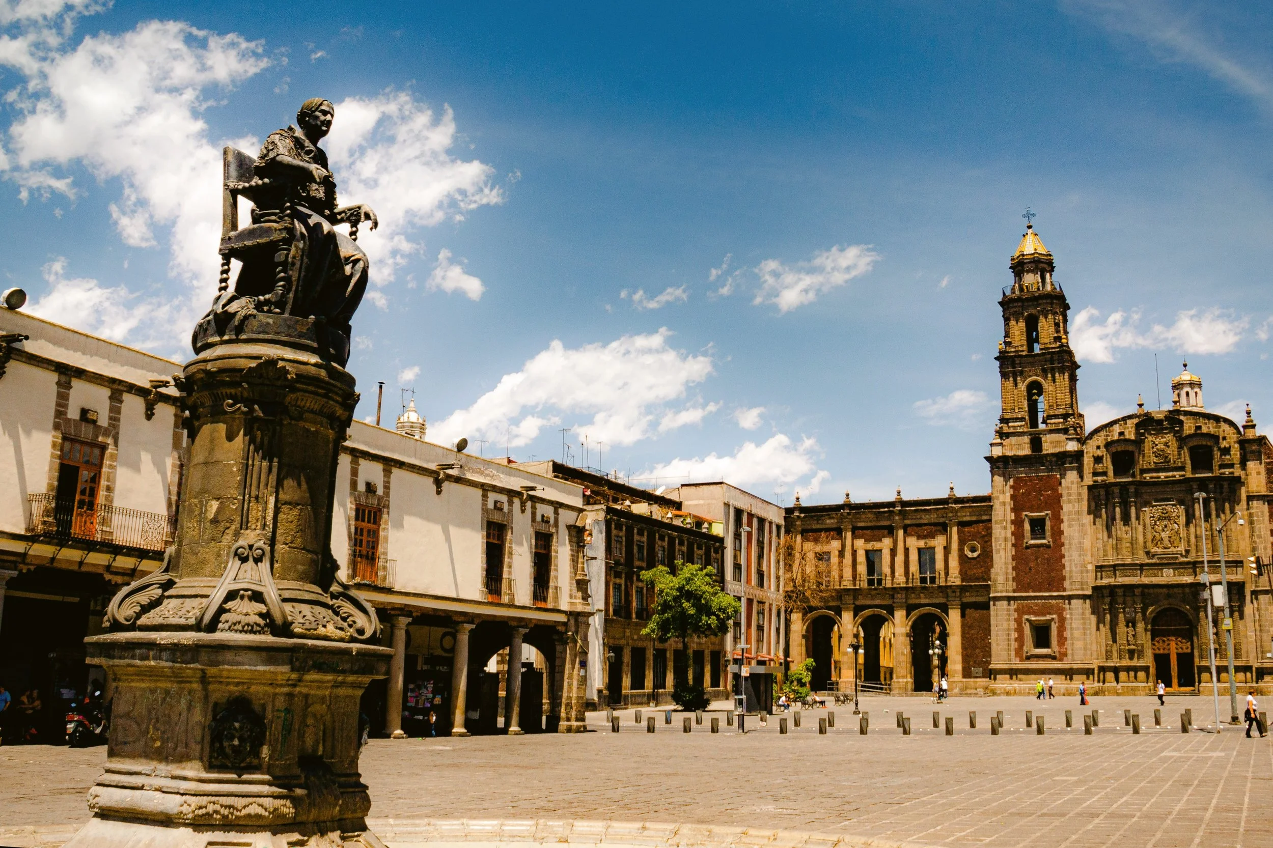 Plaza de Santo Domingo with a stone statue in the foreground and a church bell tower in the background under a blue sky, Mexico City.