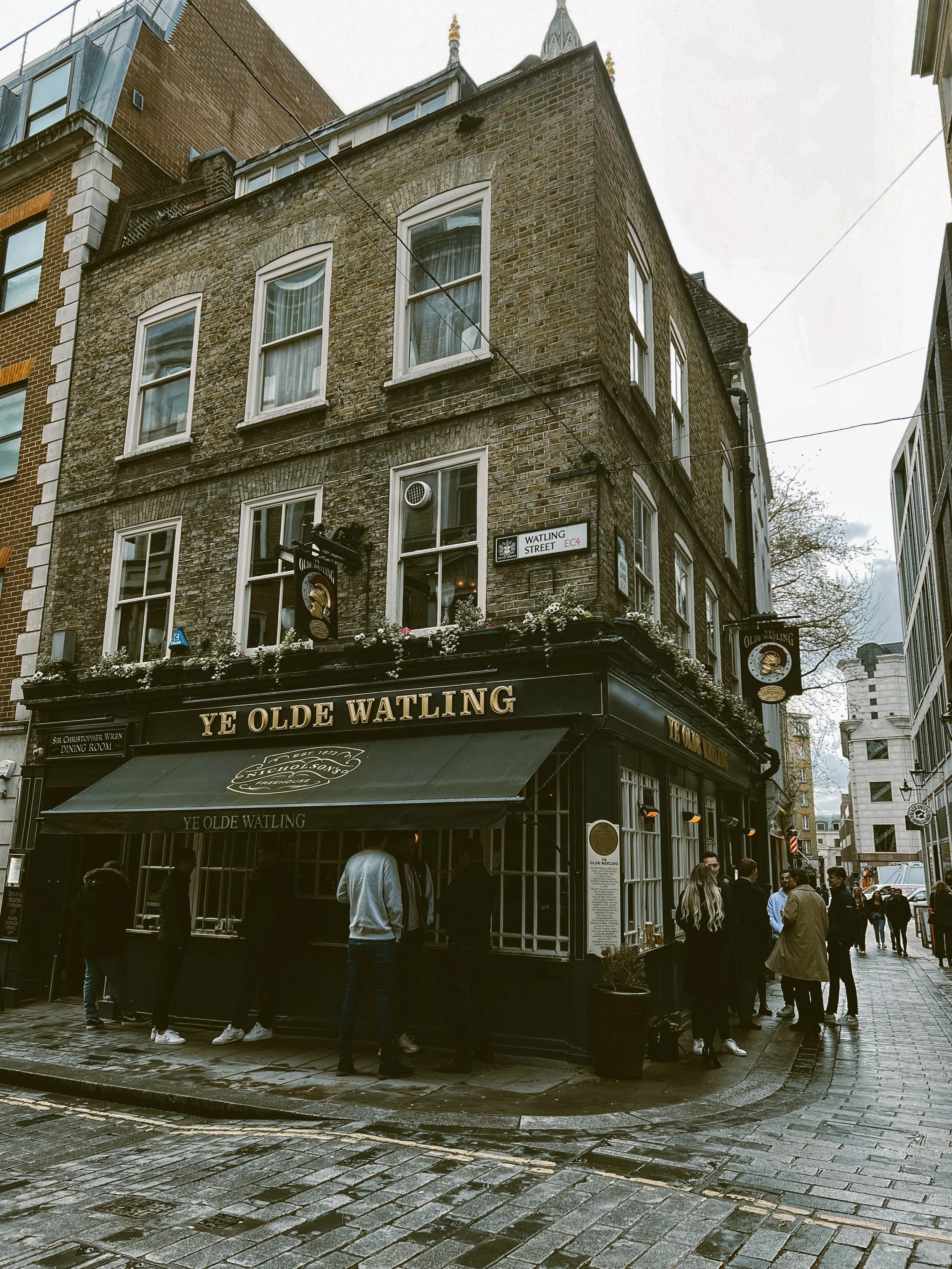 People standing outside a pub named 'Ye Olde Watling' on a rainy day, in London.