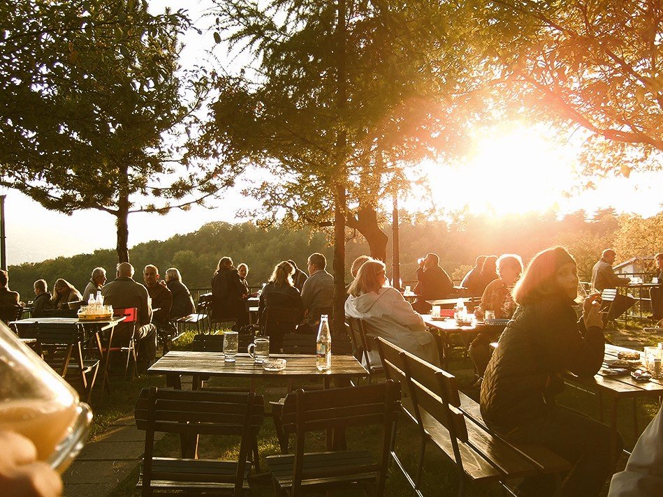 People sitting at outdoor tables enjoying the sunset in a park-like setting with trees and hills in the background.