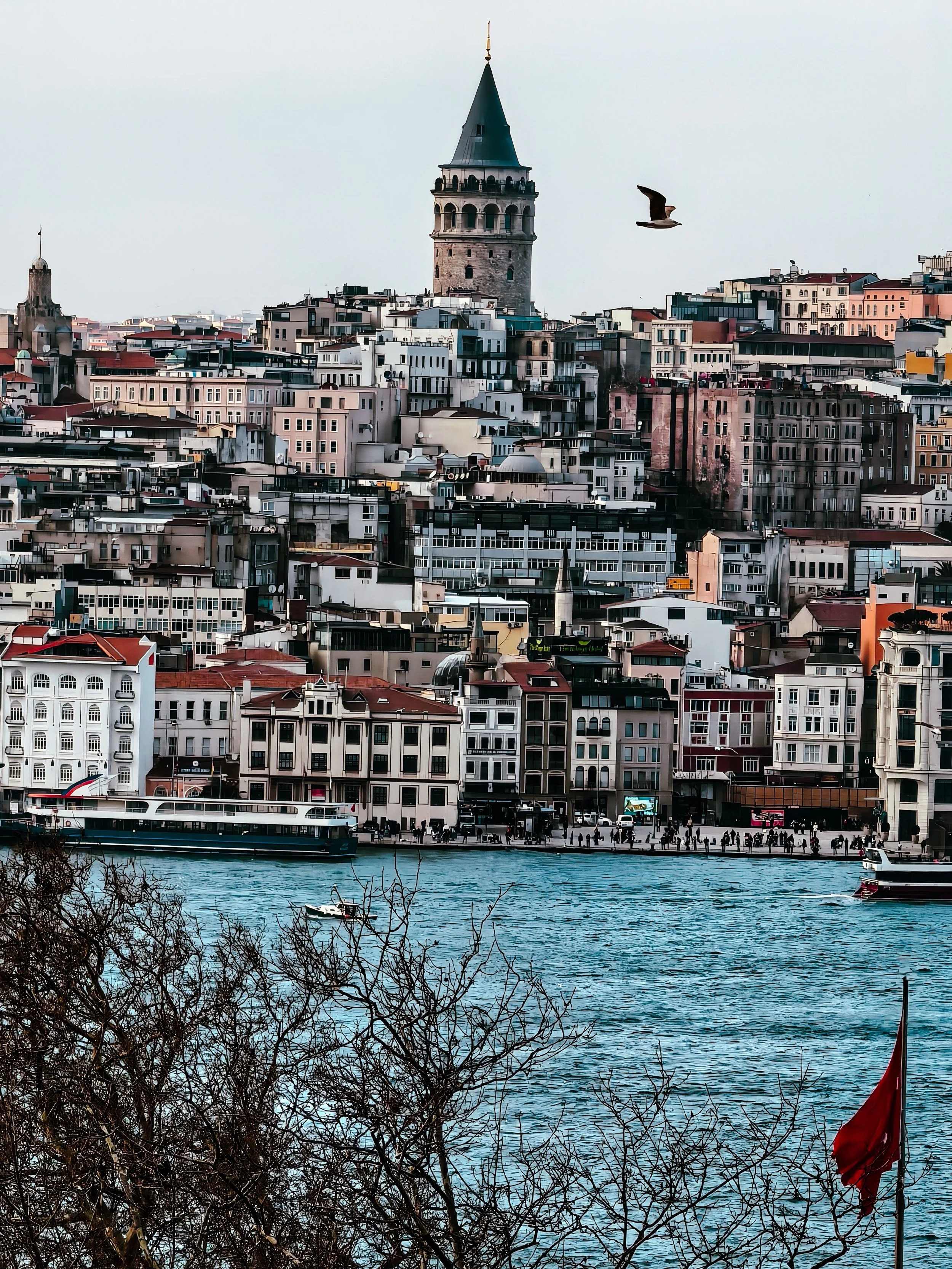 View of Istanbul skyline with the Galata Tower, waterway with boats, and buildings with a red flag in the foreground.