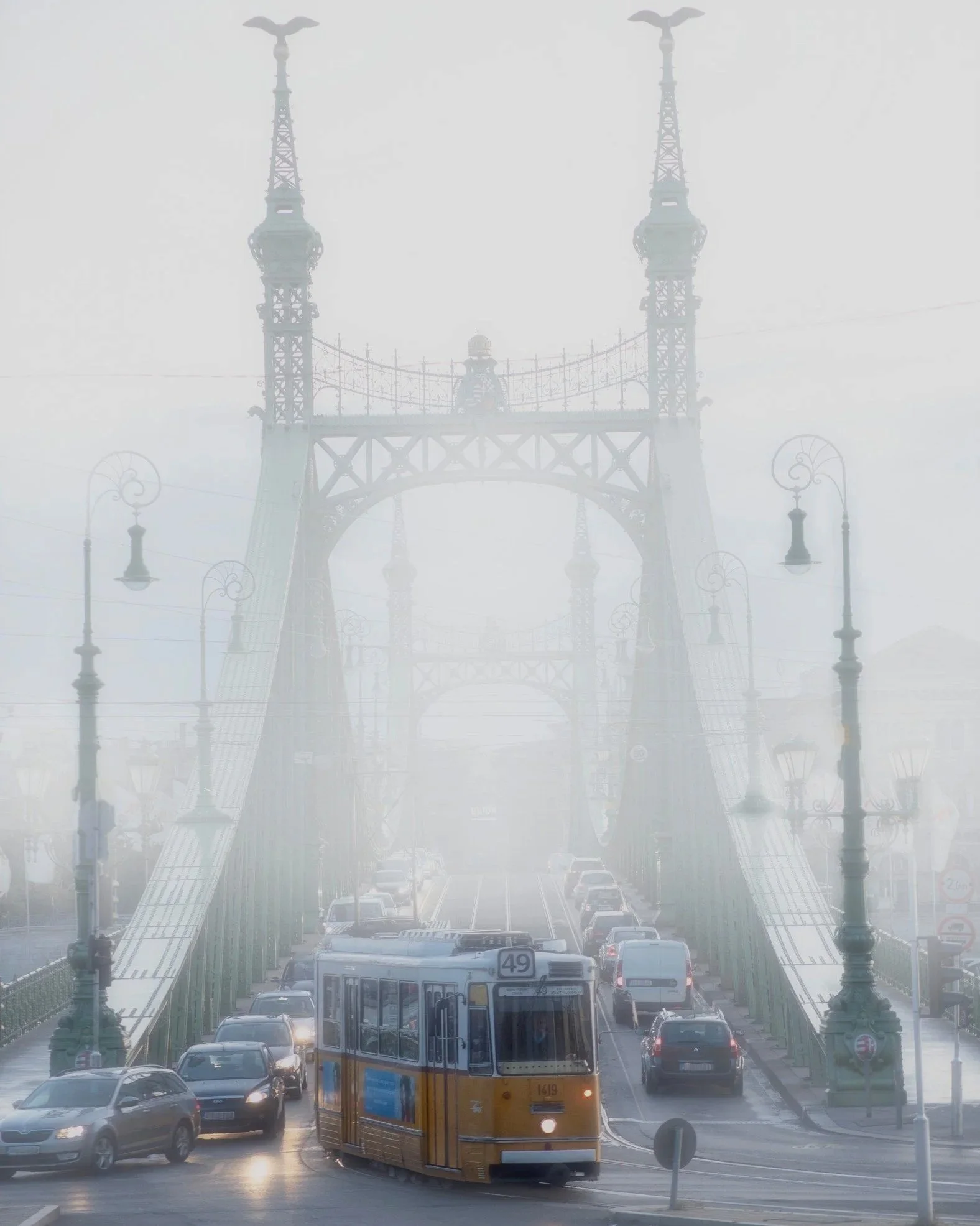Yellow tram crossing a misty Liberty Bridge (Szabadság híd) with tall green metal towers, Budapest.