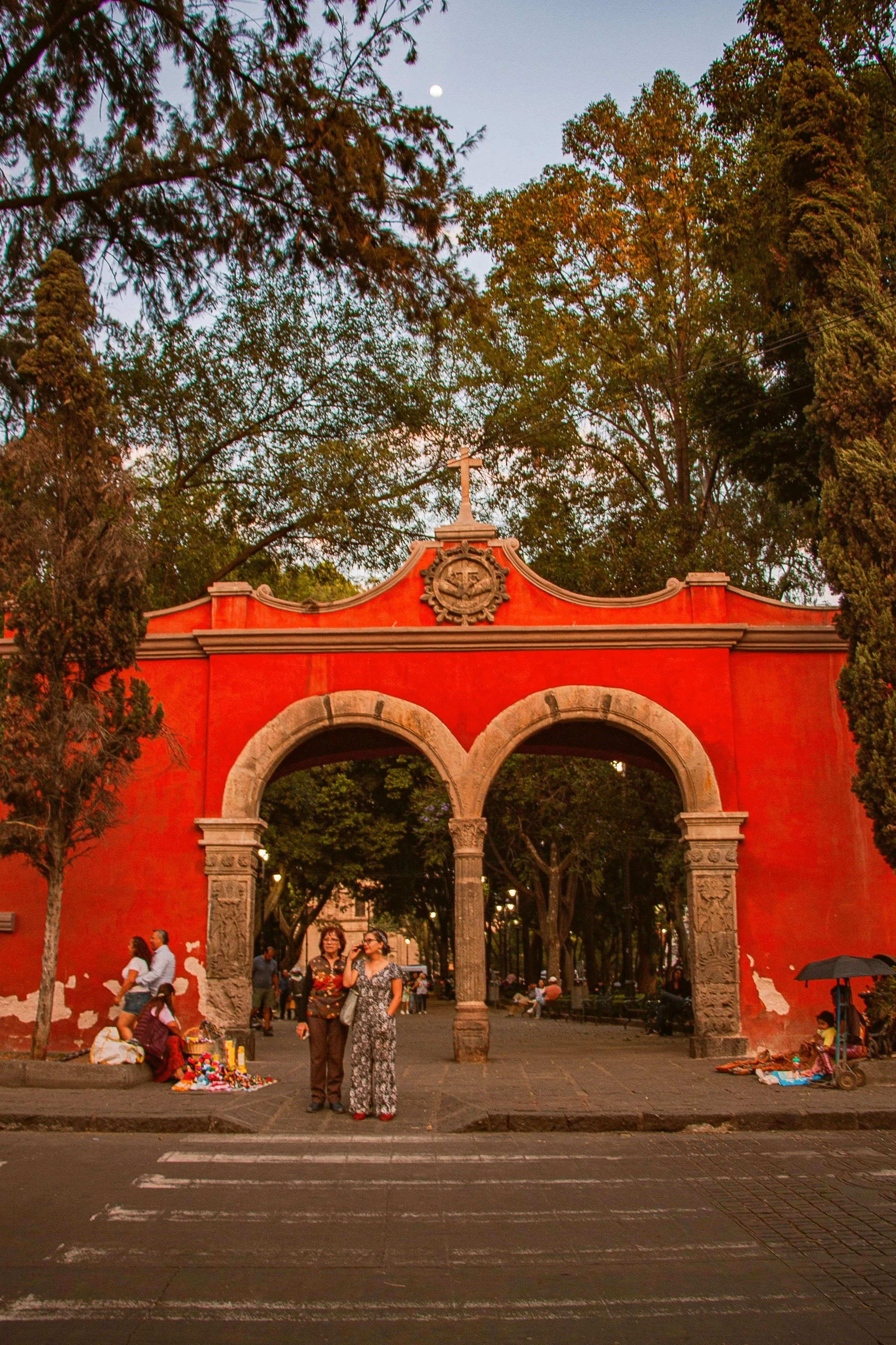 Red colonial archway with two arches and people beneath it, surrounded by trees, Coyoacán, Mexico City.