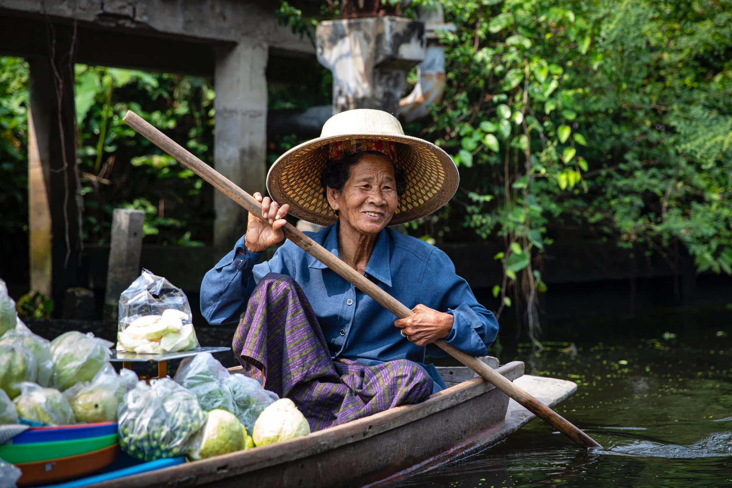 Vendor in a straw hat paddling a wooden boat loaded with fresh produce along a canal, Bangkok.
