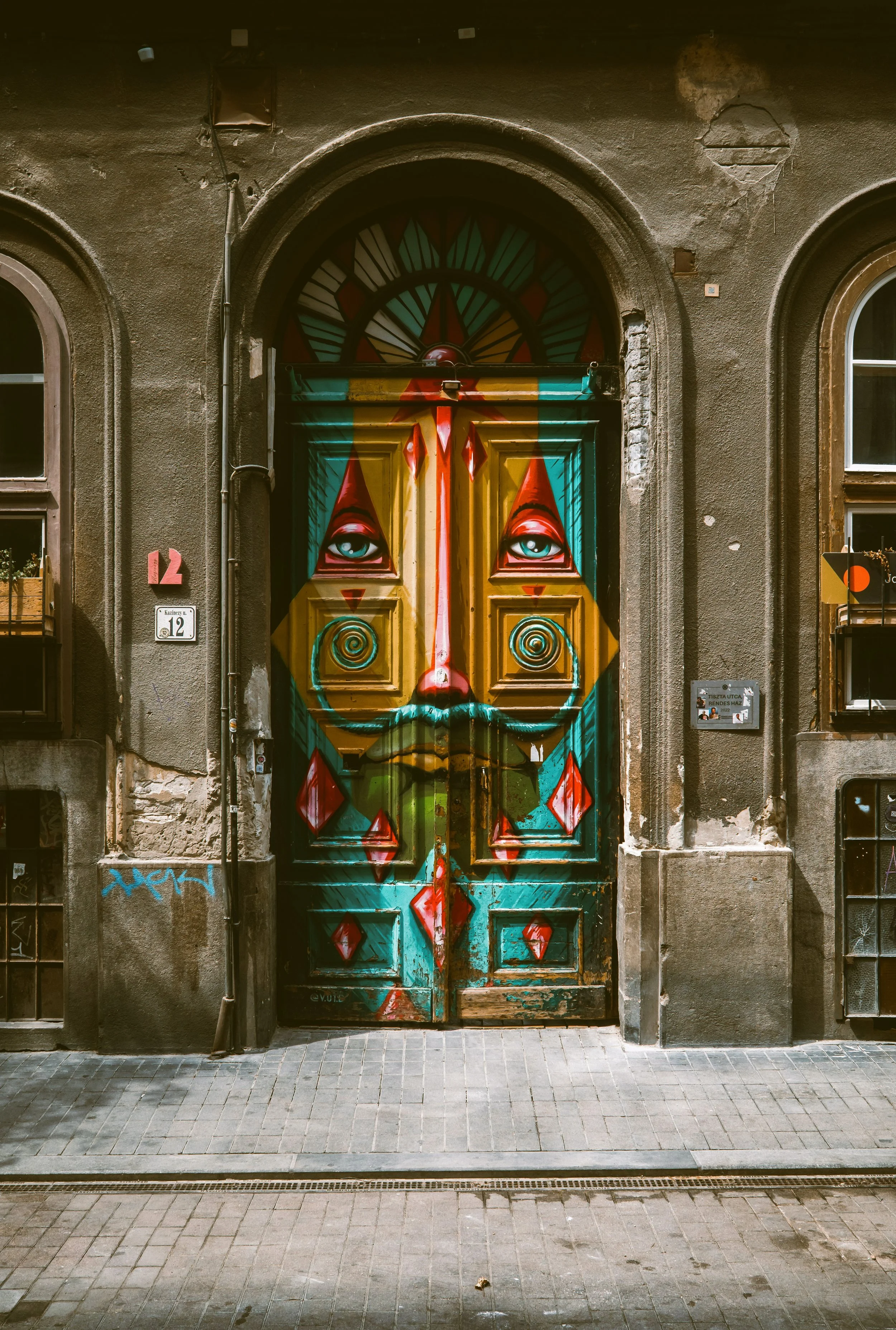 Street view of a building with a door painted as a surreal face with blue eyes, red eyebrows, a yellow nose, and mustache, in the Budapest party district.